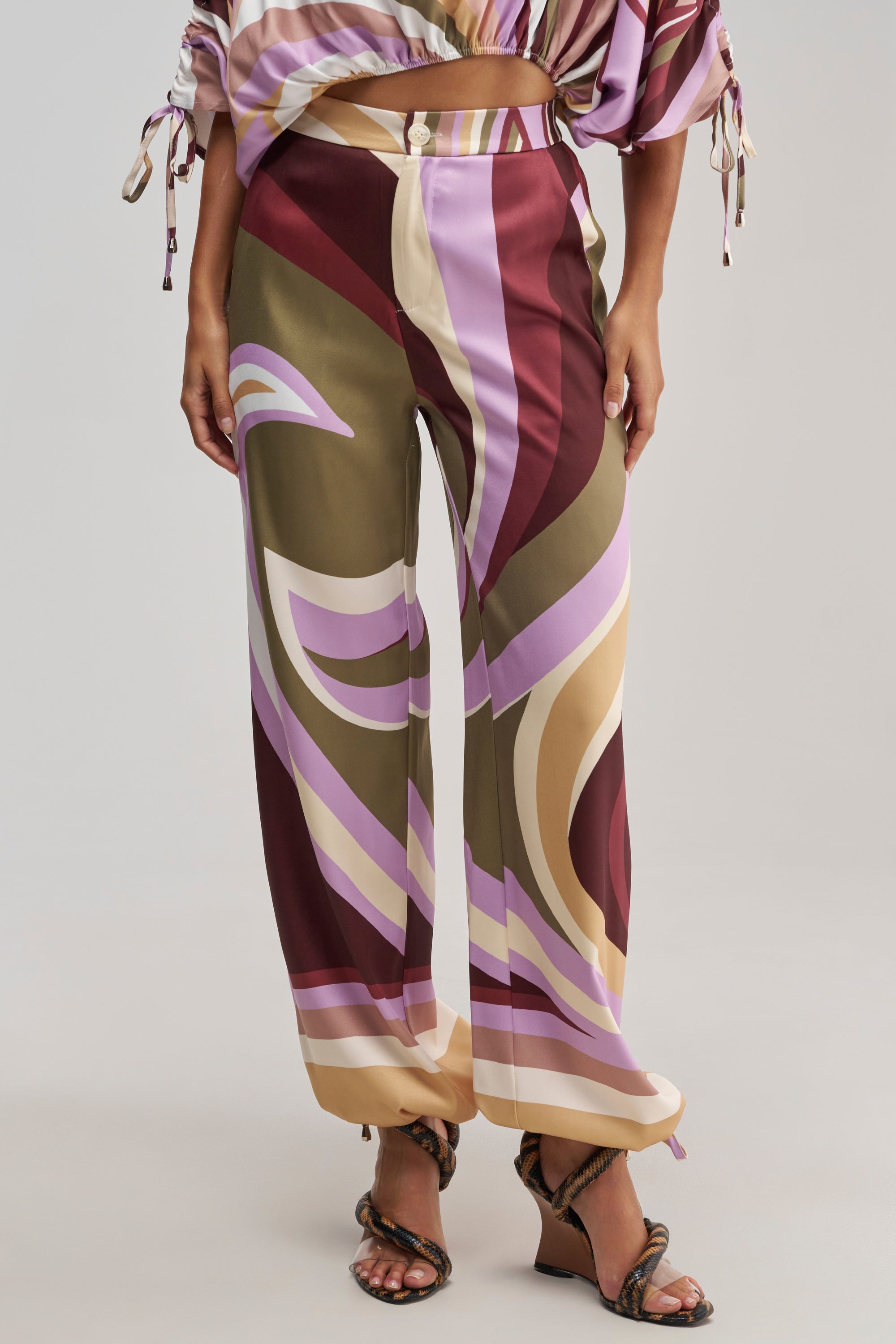 Wearing the CRAZY FOR YOU TROUSER—retro-chic, wide-leg trousers featuring a bold abstract print in maroon, mauve, brown, and beige—a person pairs them with strappy sandals and a partially visible high-rise waist top.