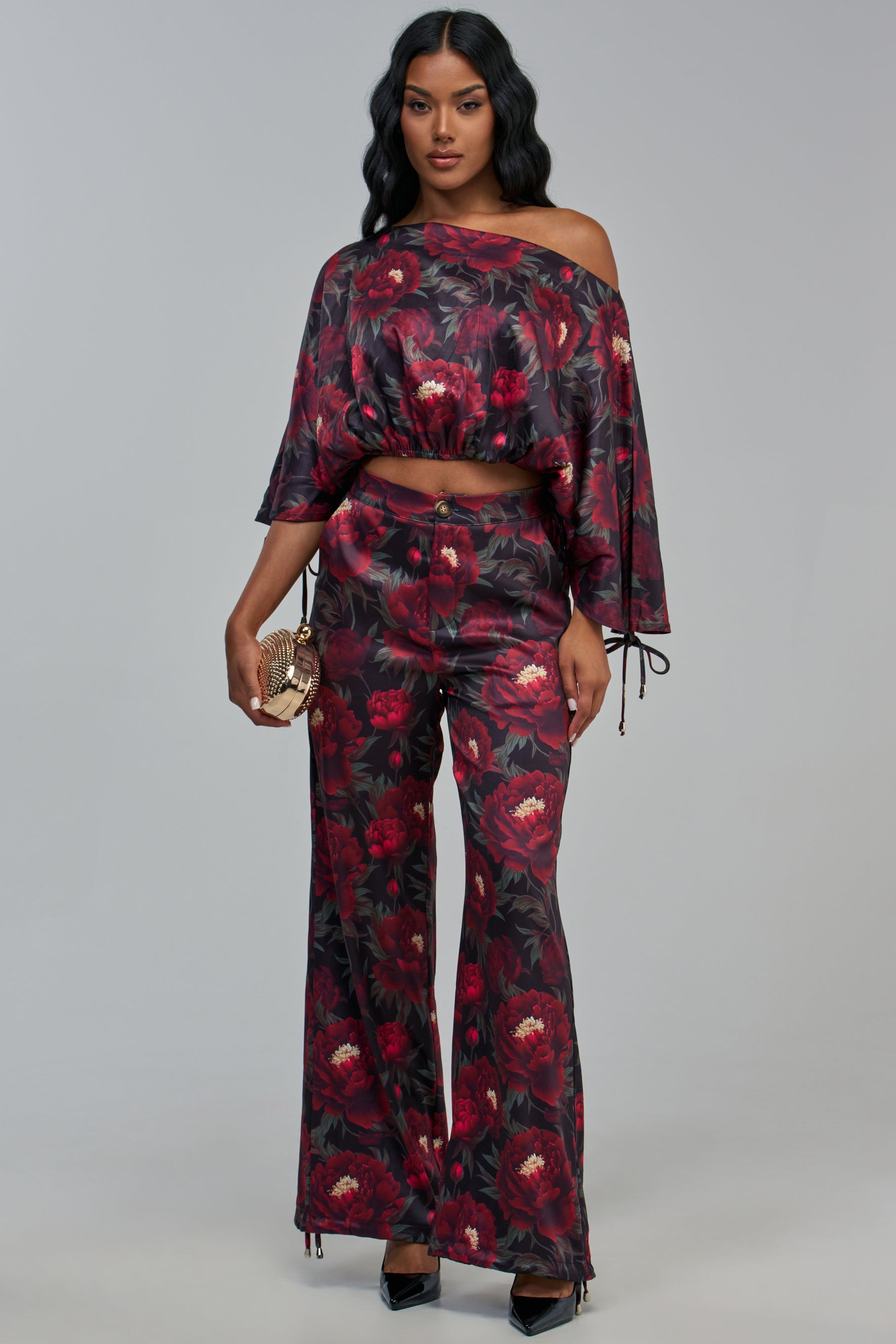 A person wears an off-the-shoulder crop top and matching high-waisted, wide-leg RARE GEM TROUSER, both featuring a red floral print with peonies.