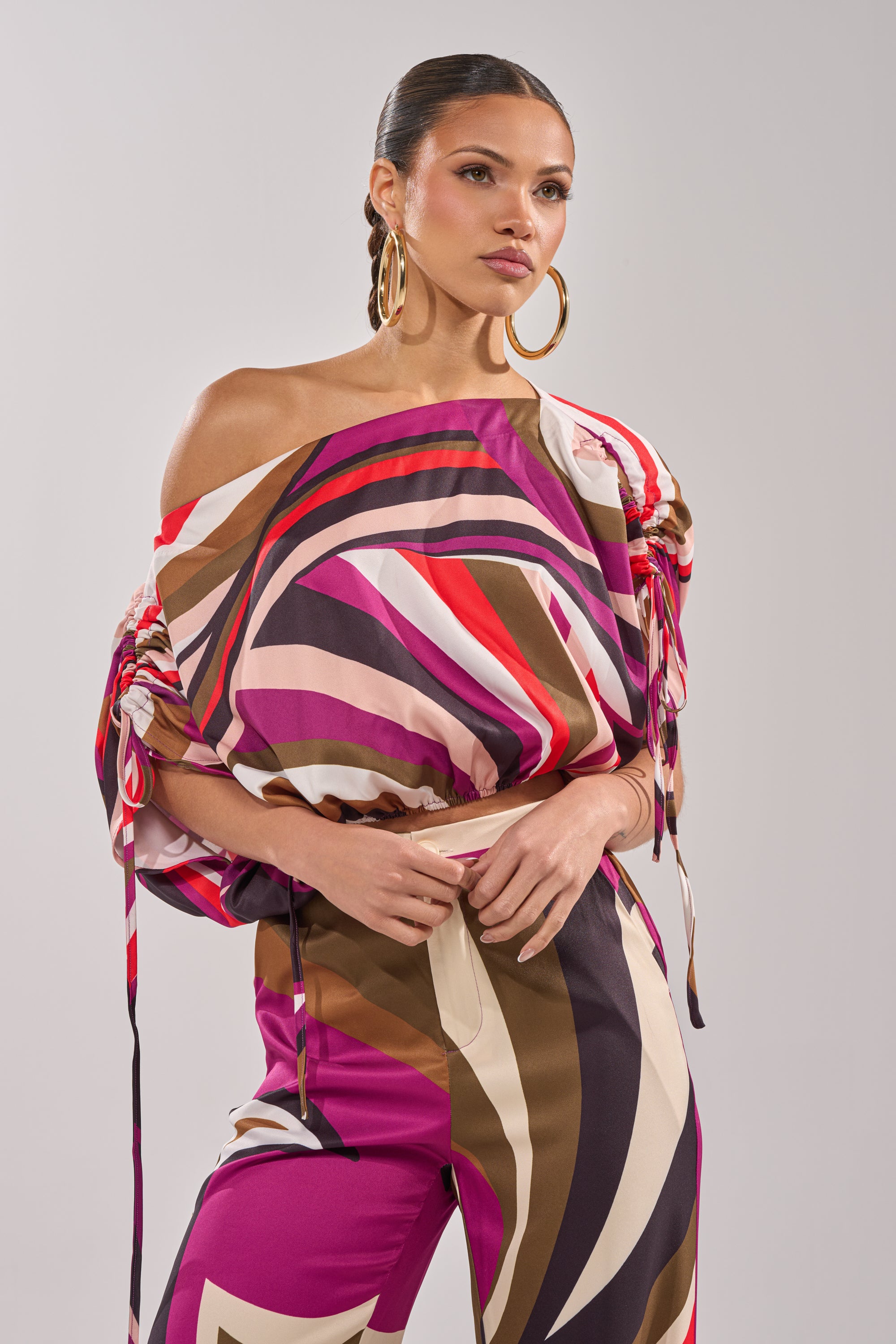 A woman models the CRAZY IN LOVE OFF THE SHOULDER BLOUSE, featuring vibrant retro geometric prints in pink, brown, and cream. She accessorizes with large hoop earrings and has sleek hair pulled back, exuding confidence. 1