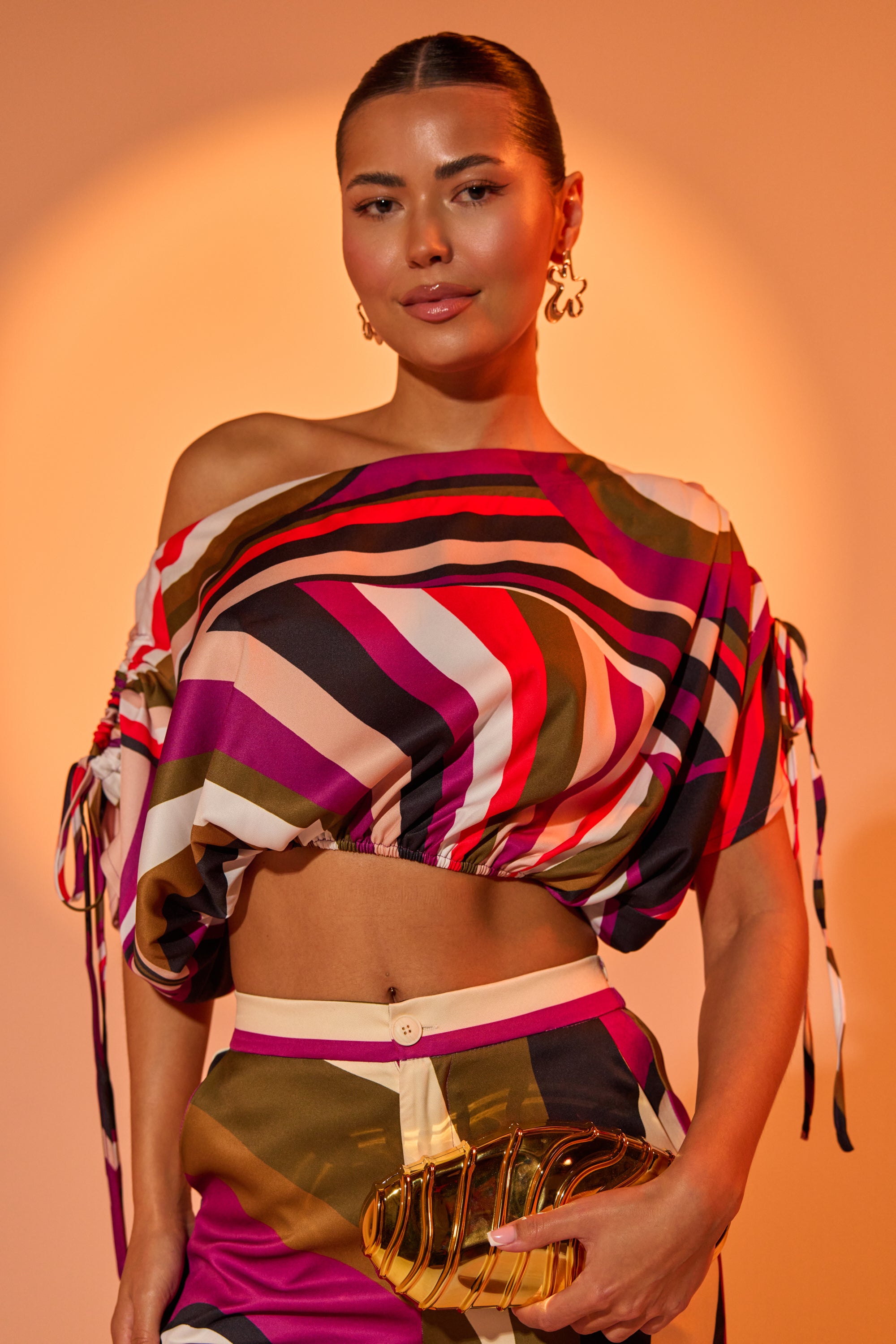 A woman poses confidently in the CRAZY IN LOVE OFF THE SHOULDER BLOUSE with voluminous sleeves, paired with matching pants and a gold clutch, her hair slicked back against a warm, orange-lit background. 0