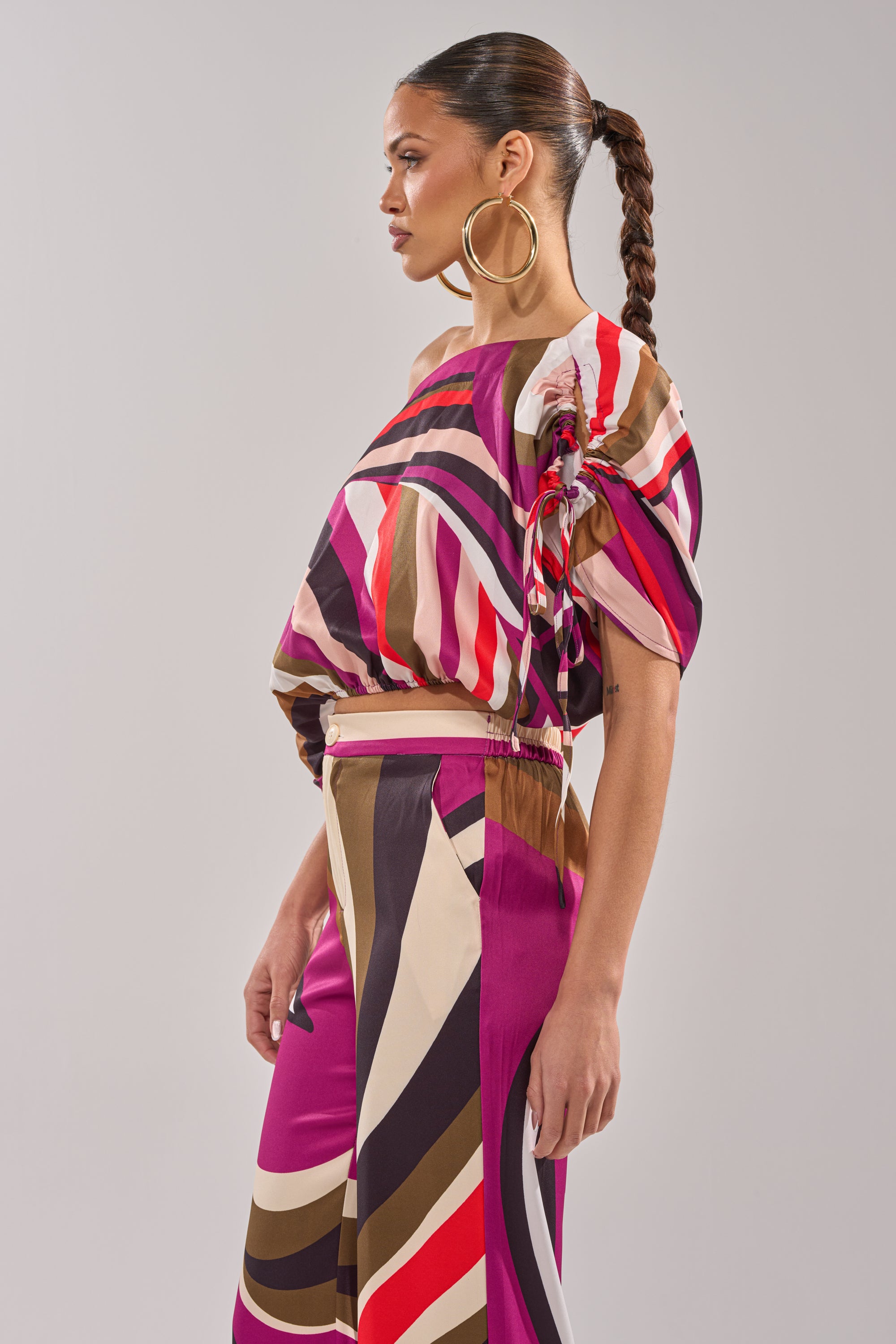 A woman with a long braided ponytail and large hoop earrings models the CRAZY IN LOVE OFF THE SHOULDER BLOUSE, featuring colorful pink, brown, cream, and red stripes. She stands against a plain background, gazing to the side. 4