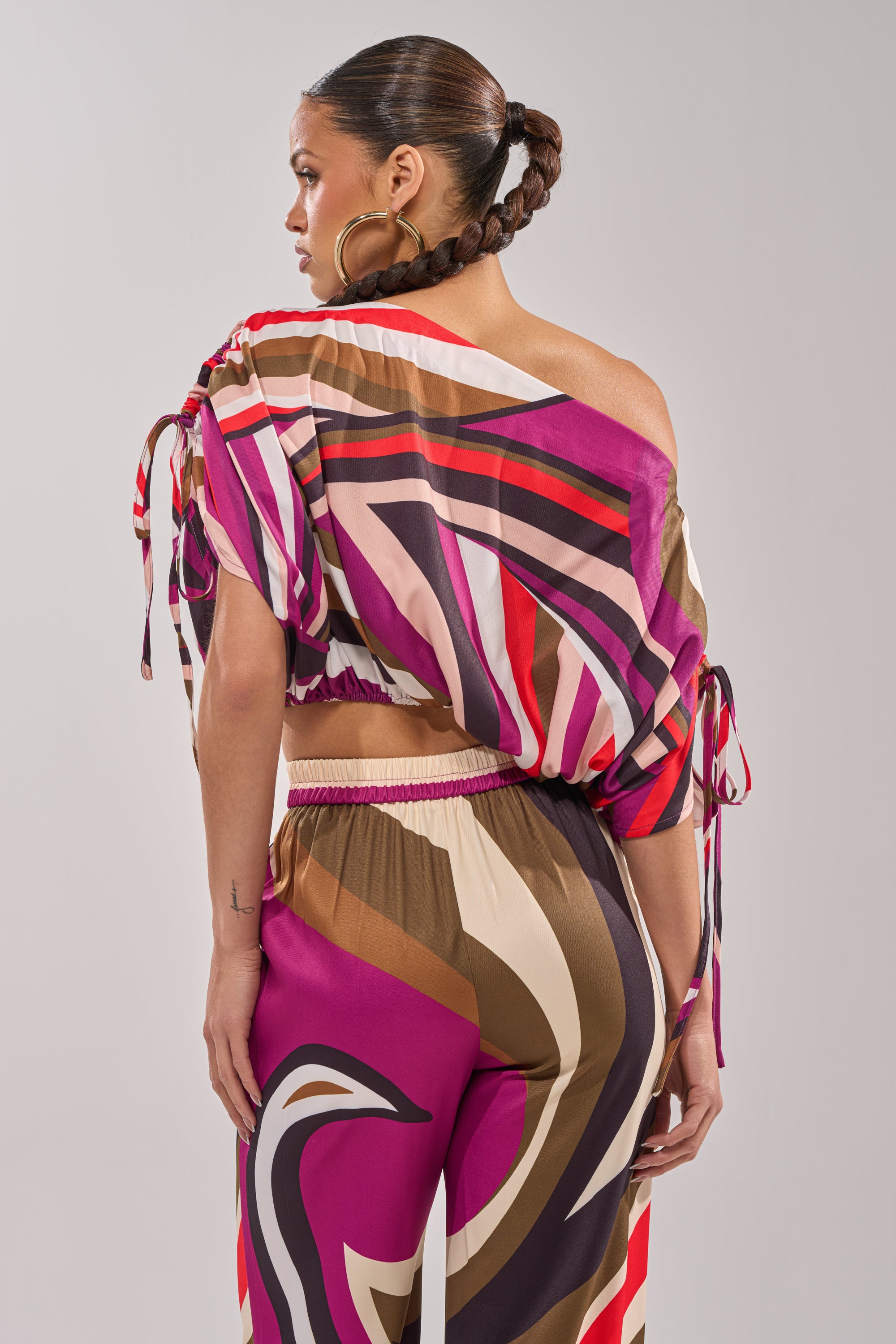 A woman with braided hair and large hoop earrings models the CRAZY IN LOVE OFF THE SHOULDER BLOUSE, a retro-chic printed top with dolman sleeves in pink, brown, and cream, paired with matching pants, standing with her back to the camera. 6