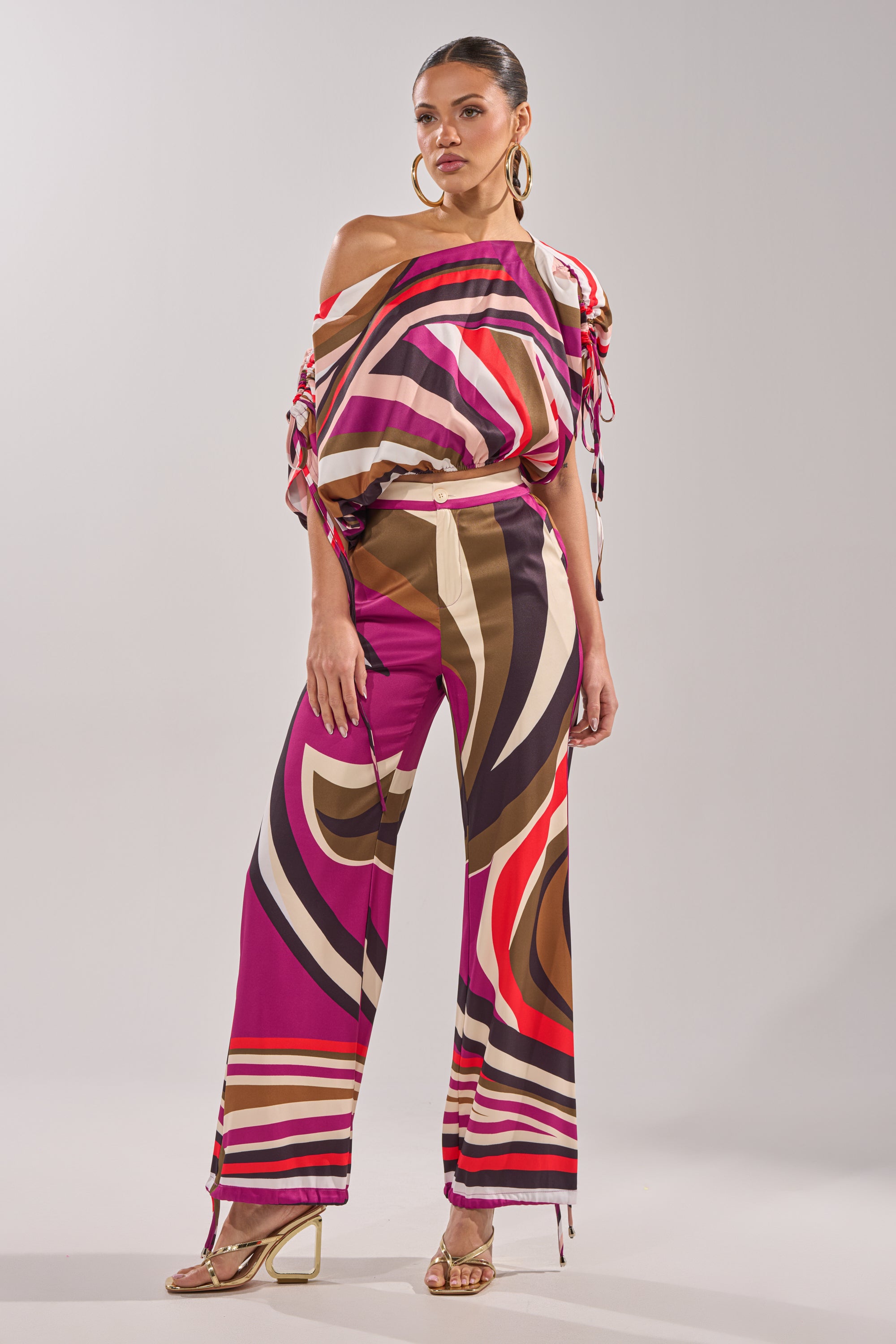 A woman models the CRAZY IN LOVE OFF THE SHOULDER BLOUSE, featuring a vibrant retro print and paired with matching wide-leg pants, gold hoop earrings, and gold high-heeled sandals against a plain light gray background. 7