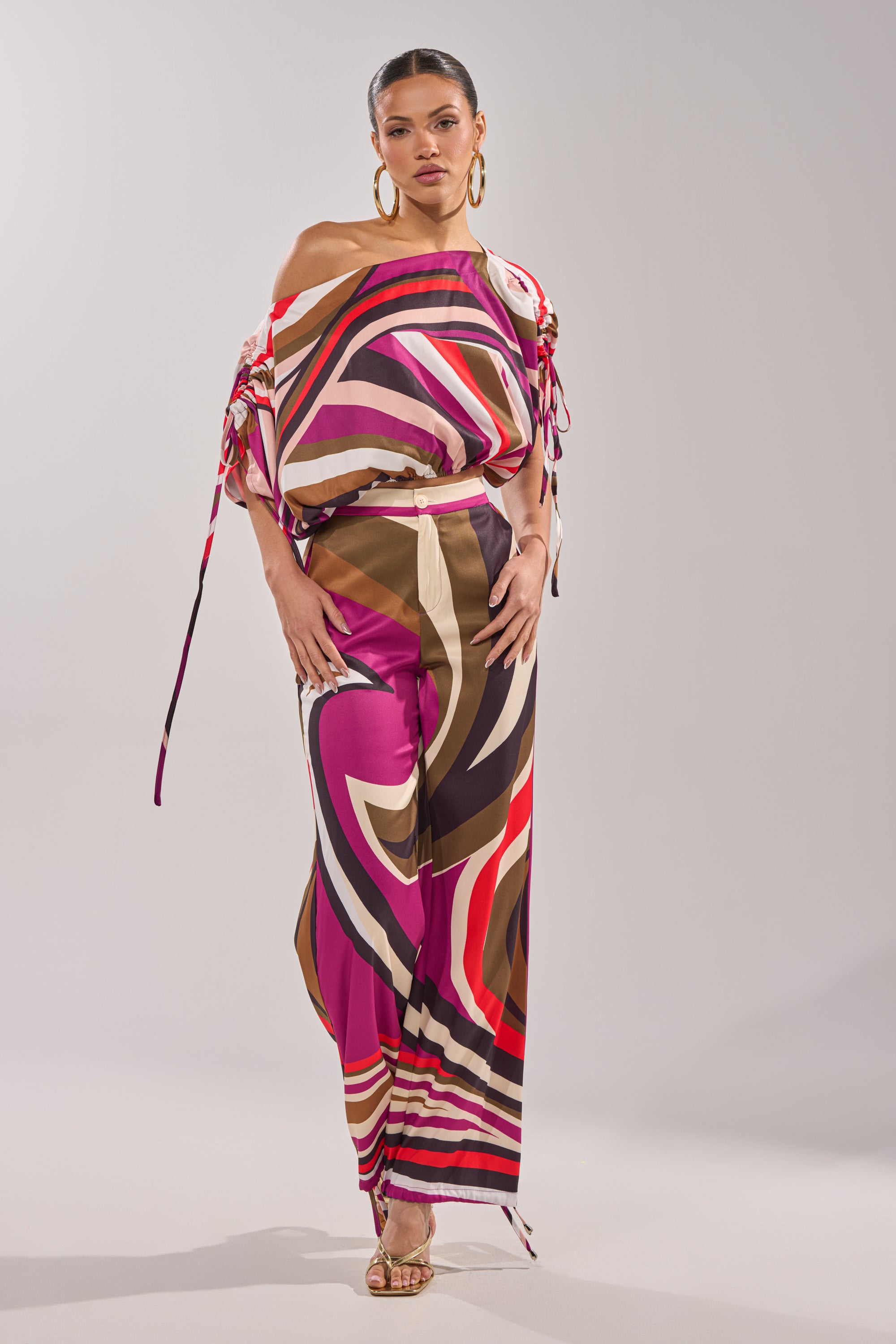 A woman poses in the CRAZY IN LOVE OFF THE SHOULDER BLOUSE with bold retro swirls of pink, red, brown, and cream, styled with hoop earrings and gold heels against a plain gray background. 9