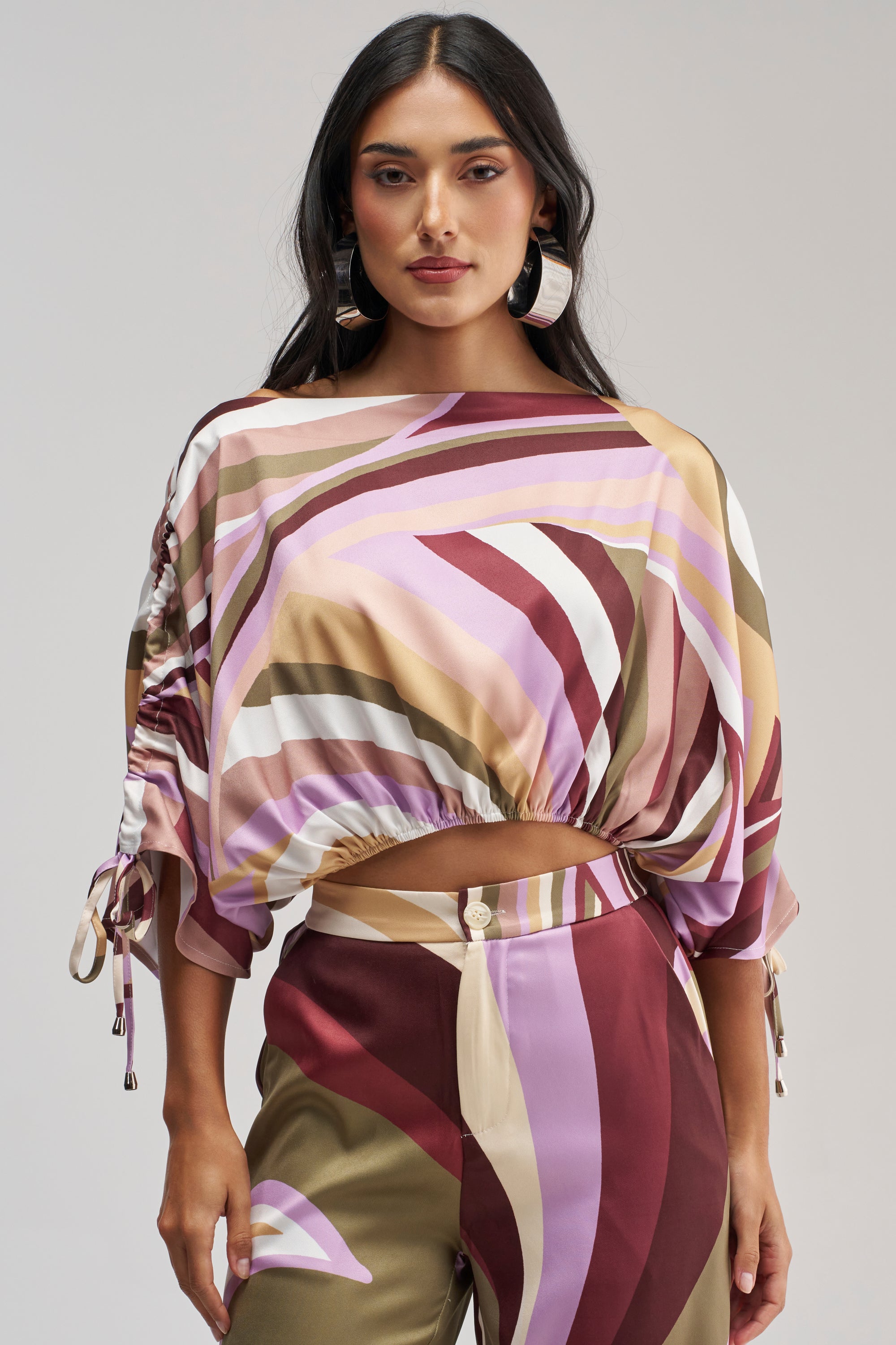 Dressed in the CRAZY FOR YOU OFF THE SHOULDER BLOUSE, the woman showcases a retro-chic look with its abstract print in shades of pink, brown, and cream. With long dark hair, hoop earrings, and a confident expression, she faces the camera.