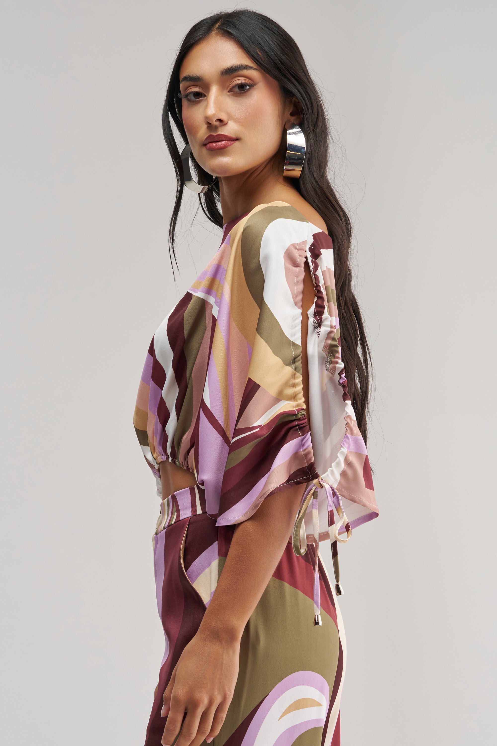A woman with long dark hair wears the CRAZY FOR YOU OFF THE SHOULDER BLOUSE, featuring a retro-chic geometric print in shades of pink, brown, and cream. She accessorizes with large hoop earrings and stands sideways against a plain background, looking confidently at the camera—her ensemble perfectly channels the abstract flair of the blouse.