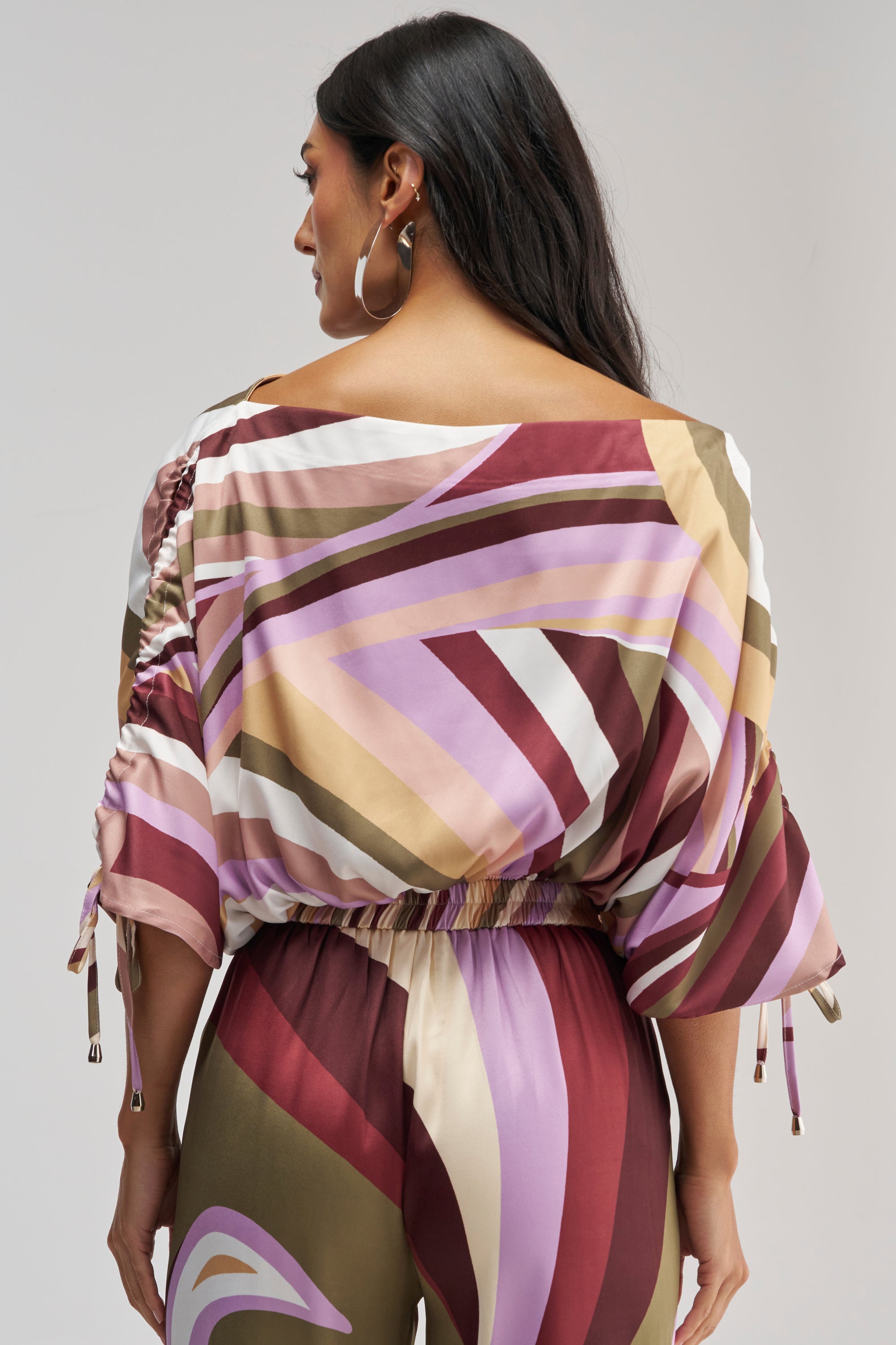 Wearing the CRAZY FOR YOU OFF THE SHOULDER BLOUSE, a woman with long dark hair faces away, highlighting the top’s flowy silhouette, ruched sleeves, and elastic waistband. The blouse features an abstract print in pink, purple, and brown tones for a retro-chic touch—perfectly paired with matching pants for a coordinated look.