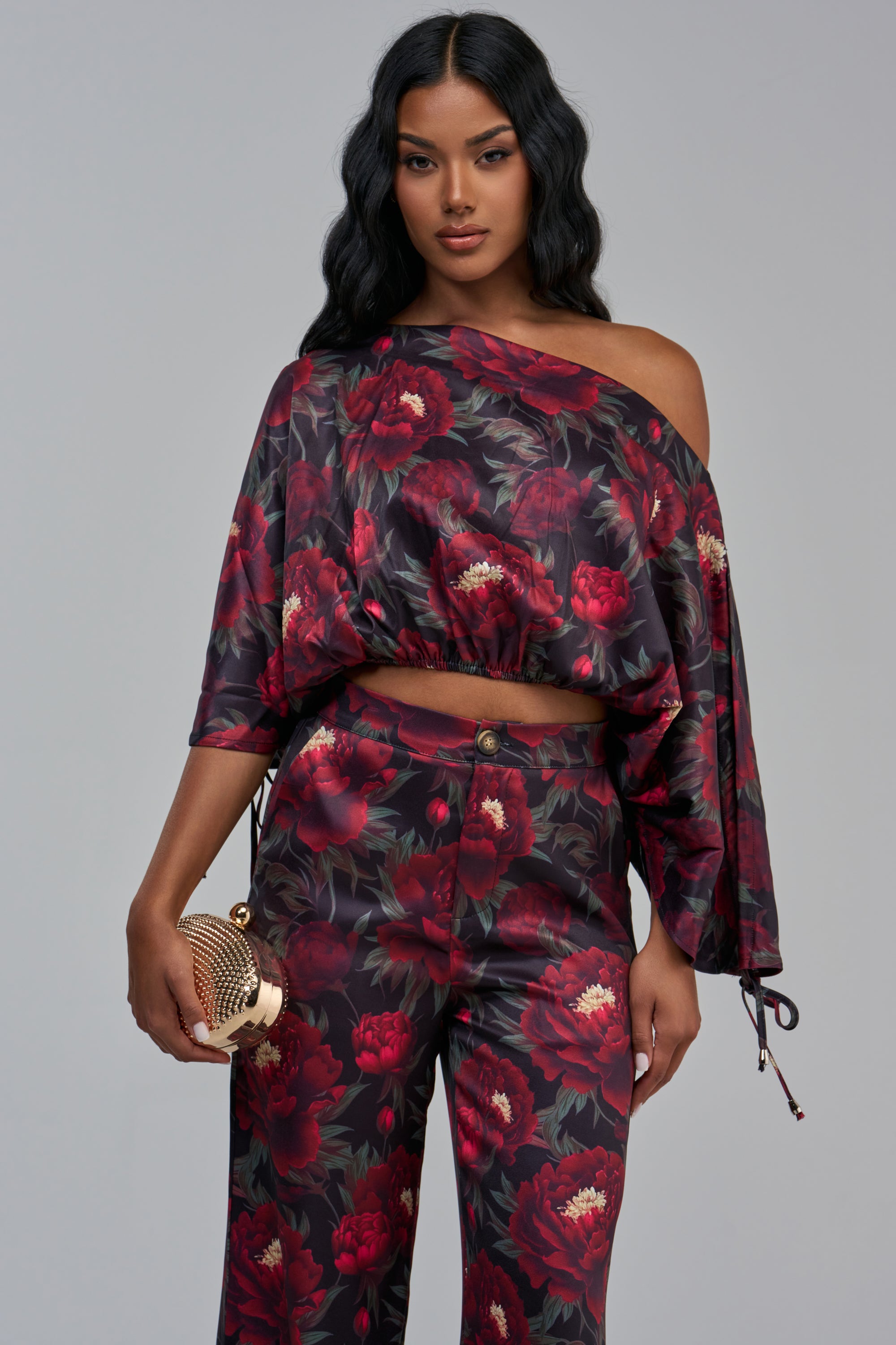 A person wearing a dark floral print RARE GEM OFF THE SHOULDER BLOUSE and matching trousers, holding a small gold textured clutch.