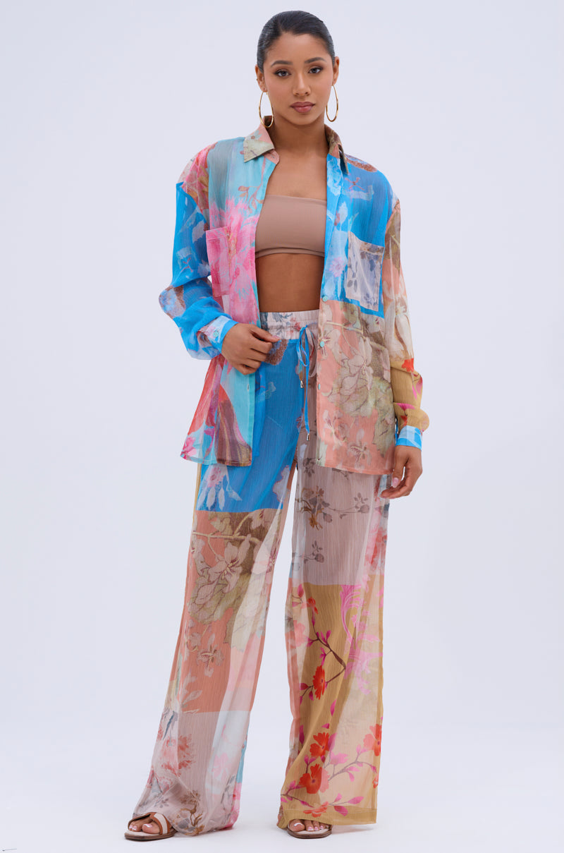 A woman wears the LILY CHIFFON WIDE LEG PANT and matching floral shirt over a beige bandeau top with sandals. She accessorizes with hoop earrings and slicked-back hair, posing against a plain white background.