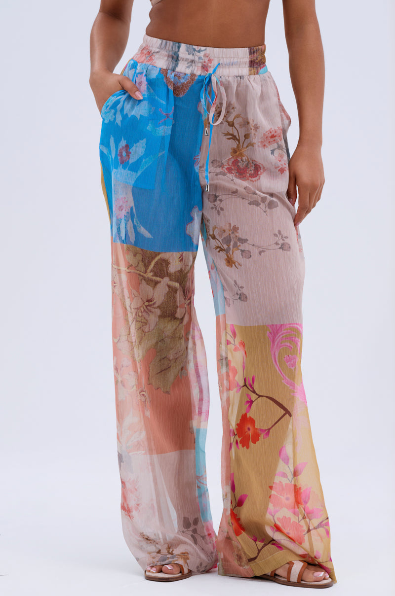 Someone wears the LILY CHIFFON WIDE LEG PANT, featuring a colorful floral patchwork, loose high-rise fit, and drawstring waist. Styled for resort wear with sandals and one hand in their pocket.