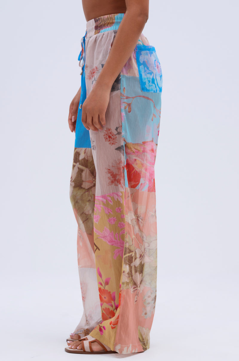A person stands sideways in the LILY CHIFFON WIDE LEG PANT, featuring a colorful patchwork floral pattern and blue pocket, paired with white sandals for a resort-ready look against a plain white background.