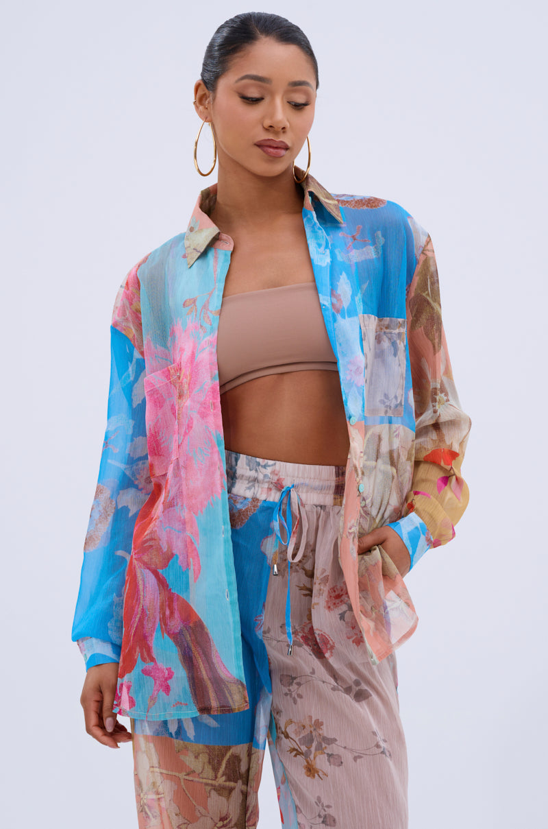 A woman poses in a resort-ready look, wearing the LILY CHIFFON BUTTON DOWN TOP over a tan bandeau and high-waisted floral beige pants, accessorized with hoop earrings and her hair tied back.