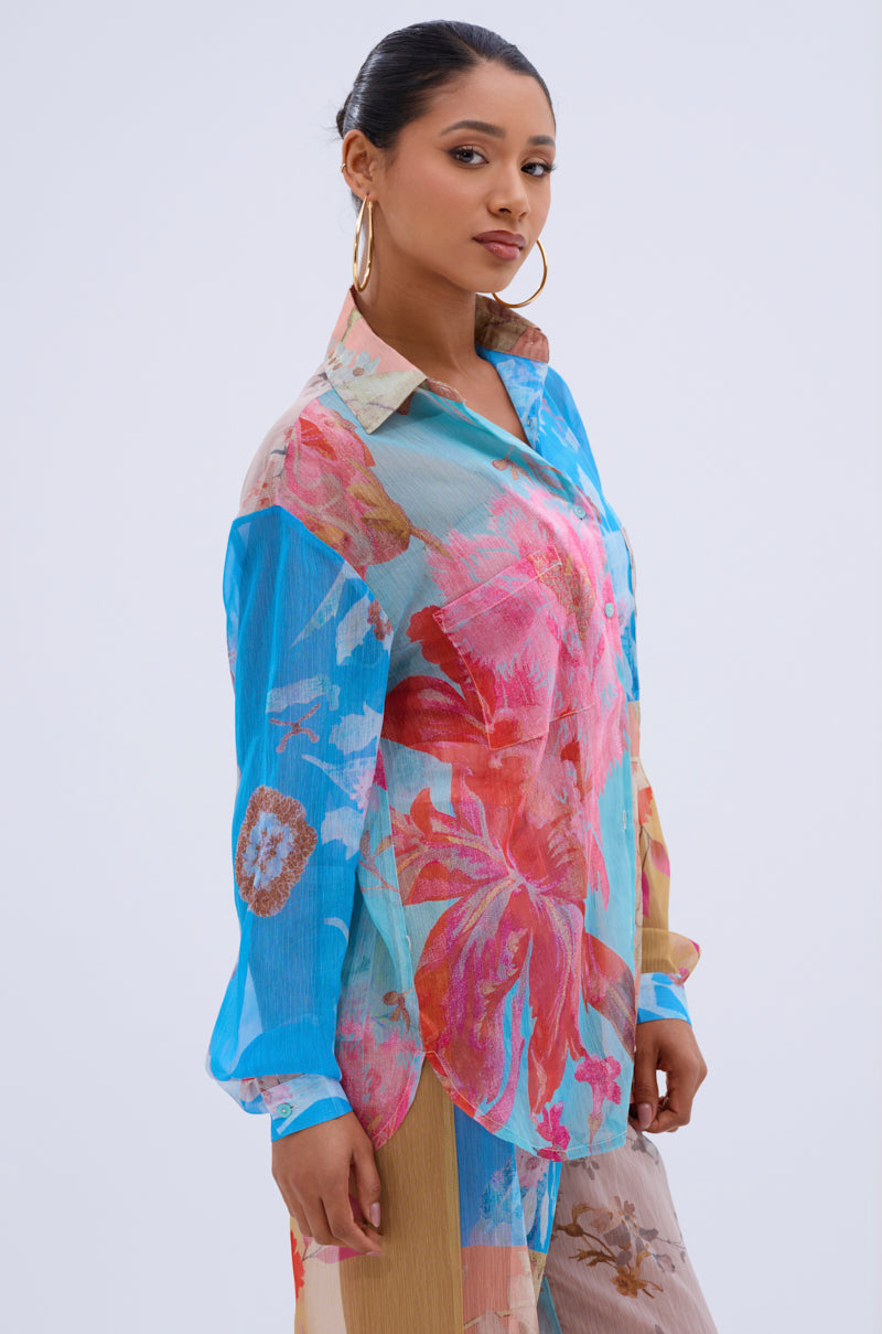 A woman models the LILY CHIFFON BUTTON DOWN TOP, a long-sleeved, floral blouse, paired with large hoop earrings and a colorful outfit, standing confidently against a light background with her hands by her sides.