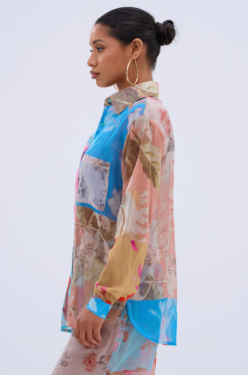 A woman with dark hair in a bun wears large hoop earrings and the LILY CHIFFON BUTTON DOWN TOP—a sheer, long-sleeve, floral blouse with a collar and chest pocket—standing in profile against a plain light background.