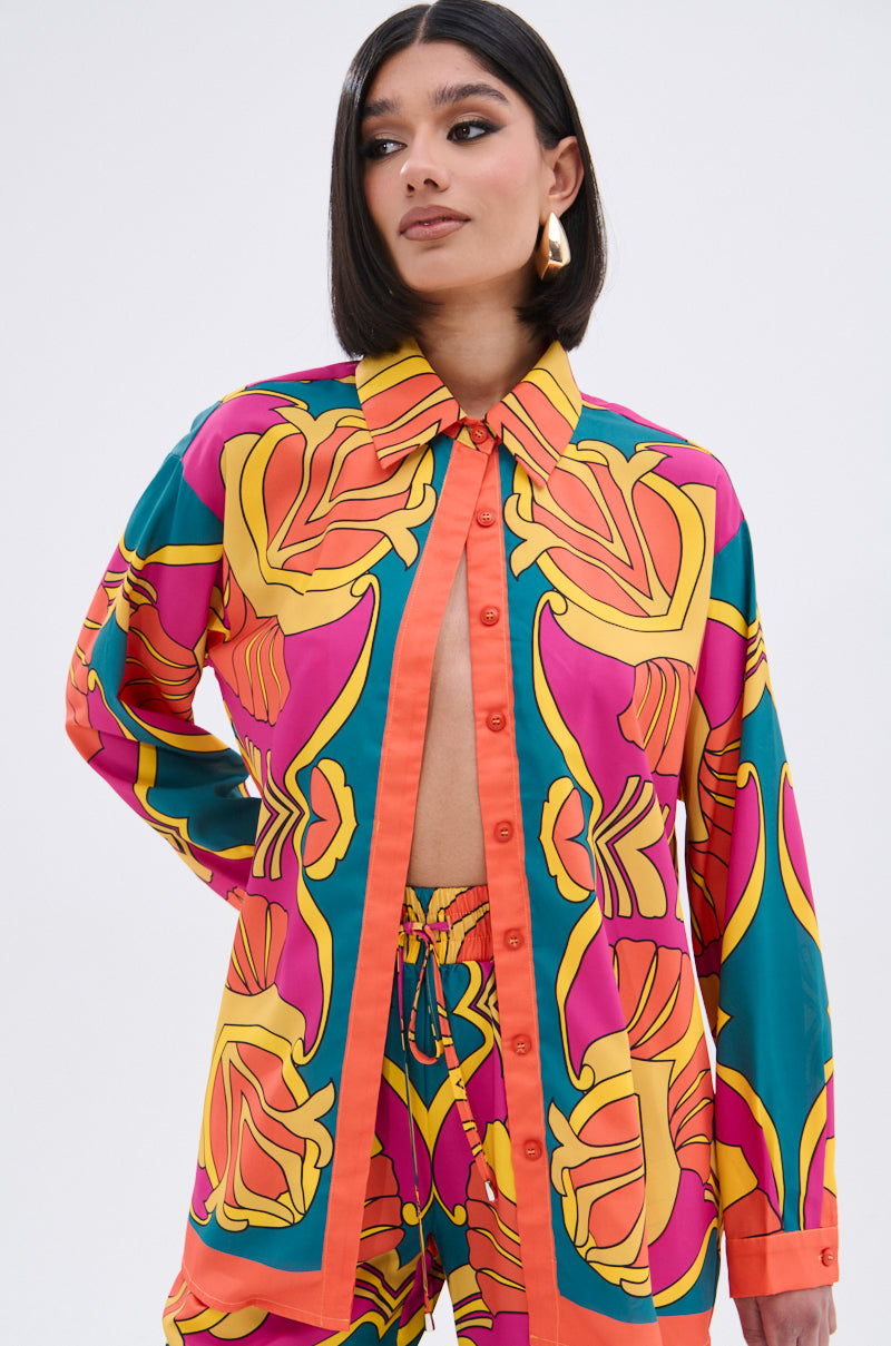 A woman with a sleek bob wears the TROPICAL DREAMS BUTTON UP SHIRT and matching shorts in vibrant pink, yellow, orange, and teal tropical print. She stands confidently with one hand on her hip, gazing off to the side.