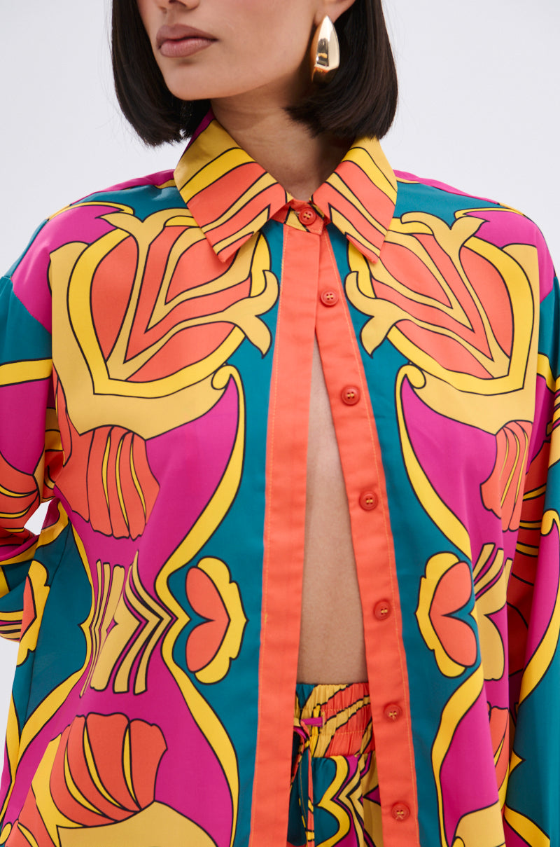 A woman with straight dark hair wears the TROPICAL DREAMS BUTTON UP SHIRT, featuring bold yellow, orange, pink, and teal tropical prints. Mostly unbuttoned to reveal a matching waistband, she accessorizes with large gold earrings.