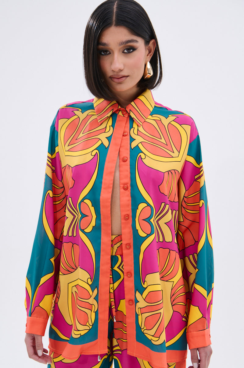 A woman with straight dark hair wears the TROPICAL DREAMS BUTTON UP SHIRT, a colorful retro top with bold orange, yellow, and magenta shapes, paired with matching pants against a plain light background.