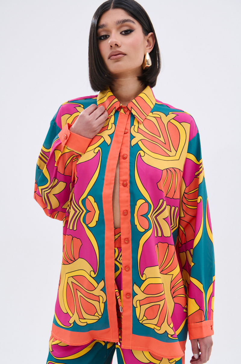 A woman with straight, shoulder-length dark hair wears the TROPICAL DREAMS BUTTON UP SHIRT, featuring a bold tropical print in pink, yellow, orange, and teal. She stands confidently against a plain background.