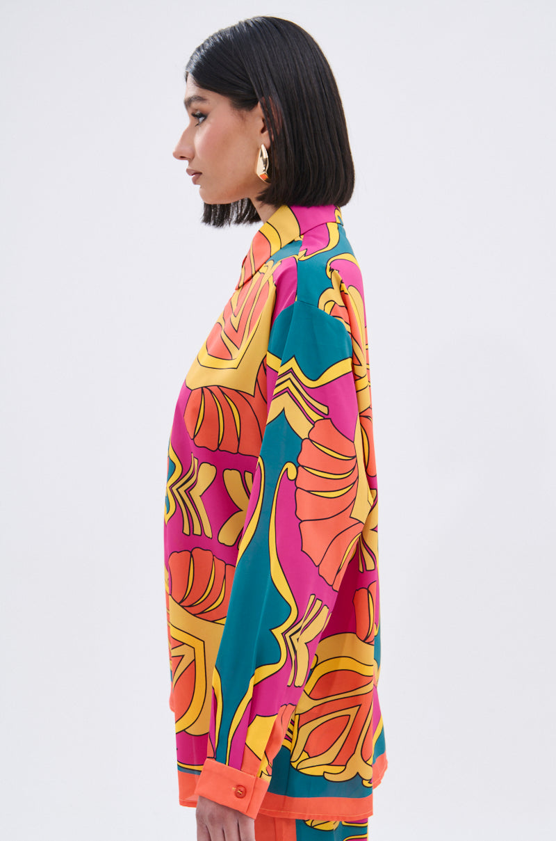 A woman with straight black hair, gold earrings, and a TROPICAL DREAMS BUTTON UP SHIRT—featuring a vibrant orange, yellow, teal, and pink tropical print—stands in profile against a plain light background.