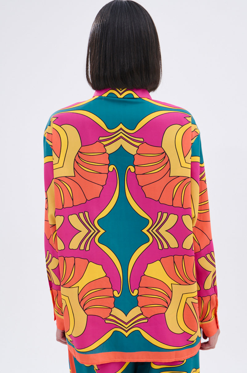 A person with straight, shoulder-length dark hair is seen from the back wearing the TROPICAL DREAMS BUTTON UP SHIRT, which features bold, symmetrical yellow, pink, orange, and teal patterns against a plain background.