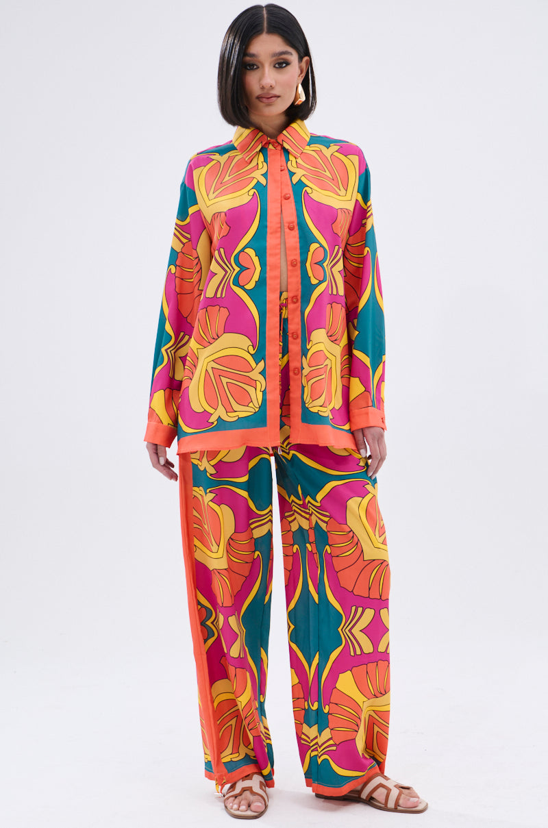 A woman wears the TROPICAL DREAMS BUTTON UP SHIRT, a colorful, loose-fit top with matching wide-leg pants in a vibrant orange, yellow, pink, and teal tropical print. She finishes the look with sandals, hoop earrings, and straight dark hair.