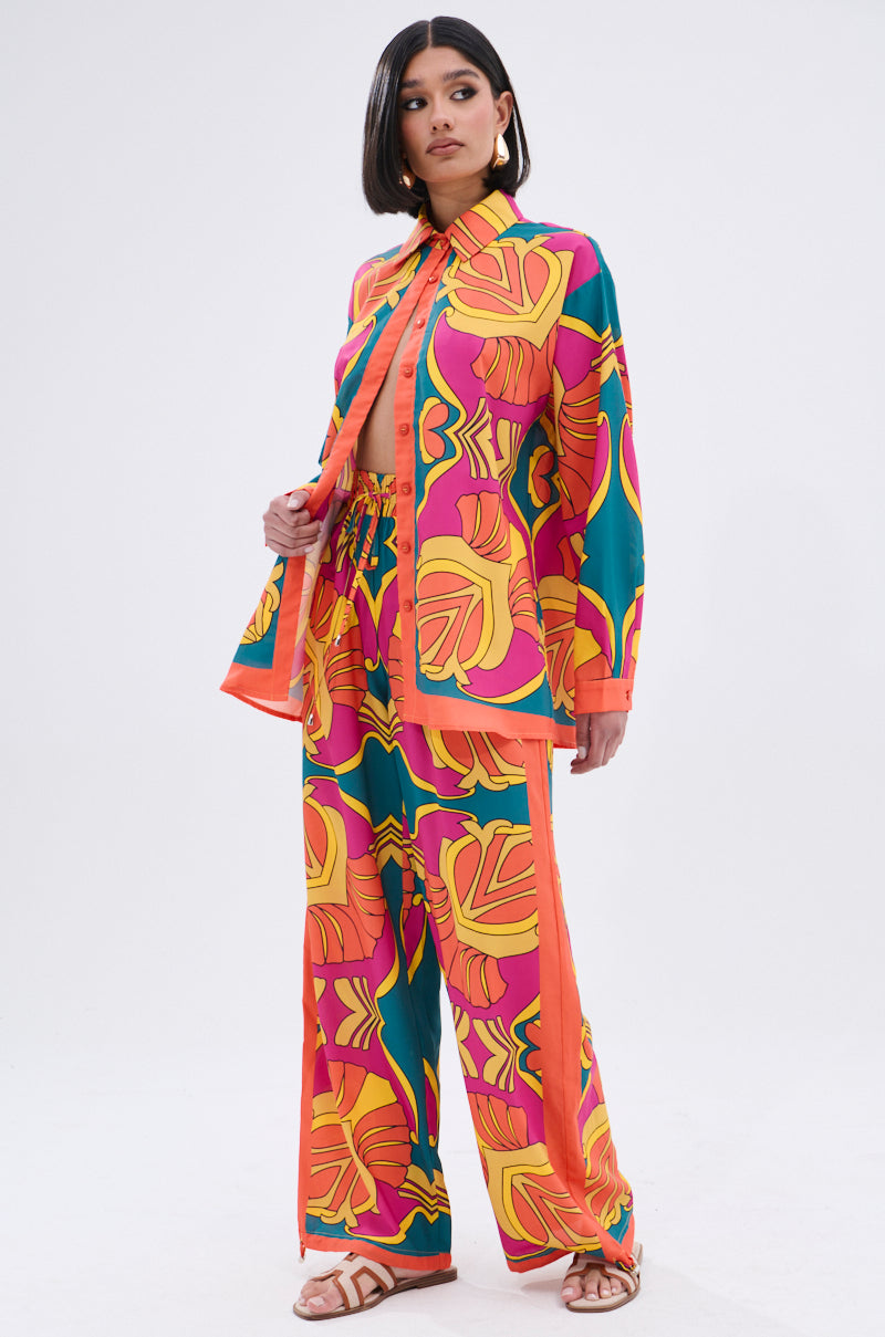 A woman stands confidently in the TROPICAL DREAMS BUTTON UP SHIRT, featuring a bold tropical print in orange, yellow, pink, and teal. She styles it with white sandals and large hoop earrings against a plain background.