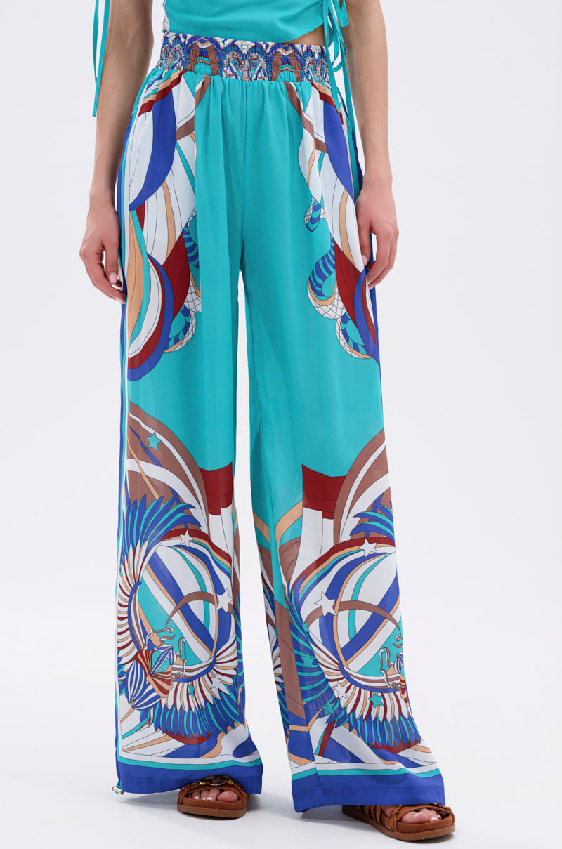The lower half of a person wearing the ASHTIN PRINTED PALAZZO PANT, featuring wide-leg, high-waisted turquoise fabric with colorful geometric and abstract motifs, paired with brown sandals—perfect resortwear inspiration. 1