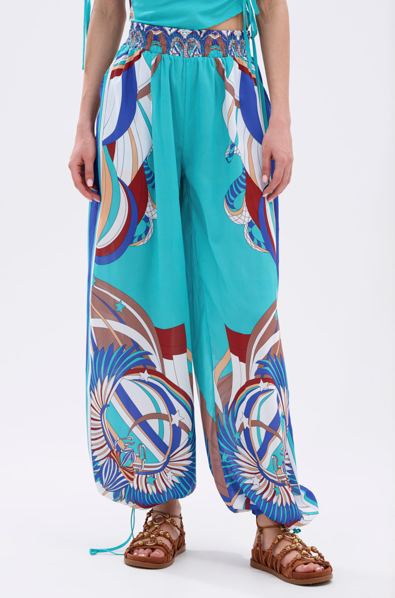 A person models the ASHTIN PRINTED PALAZZO PANT—loose turquoise pants with vibrant blue, red, and white abstract motifs—paired with brown strappy sandals for a stylish resortwear look. Their torso is partially visible, arms at their sides. 2