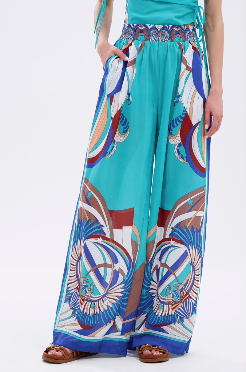 Wearing the ASHTIN PRINTED PALAZZO PANT, a wide-leg, high-waisted turquoise pant with bold geometric and feather motifs, paired with a matching top and brown sandals. Hands in pockets, they exude relaxed resortwear vibes. 3