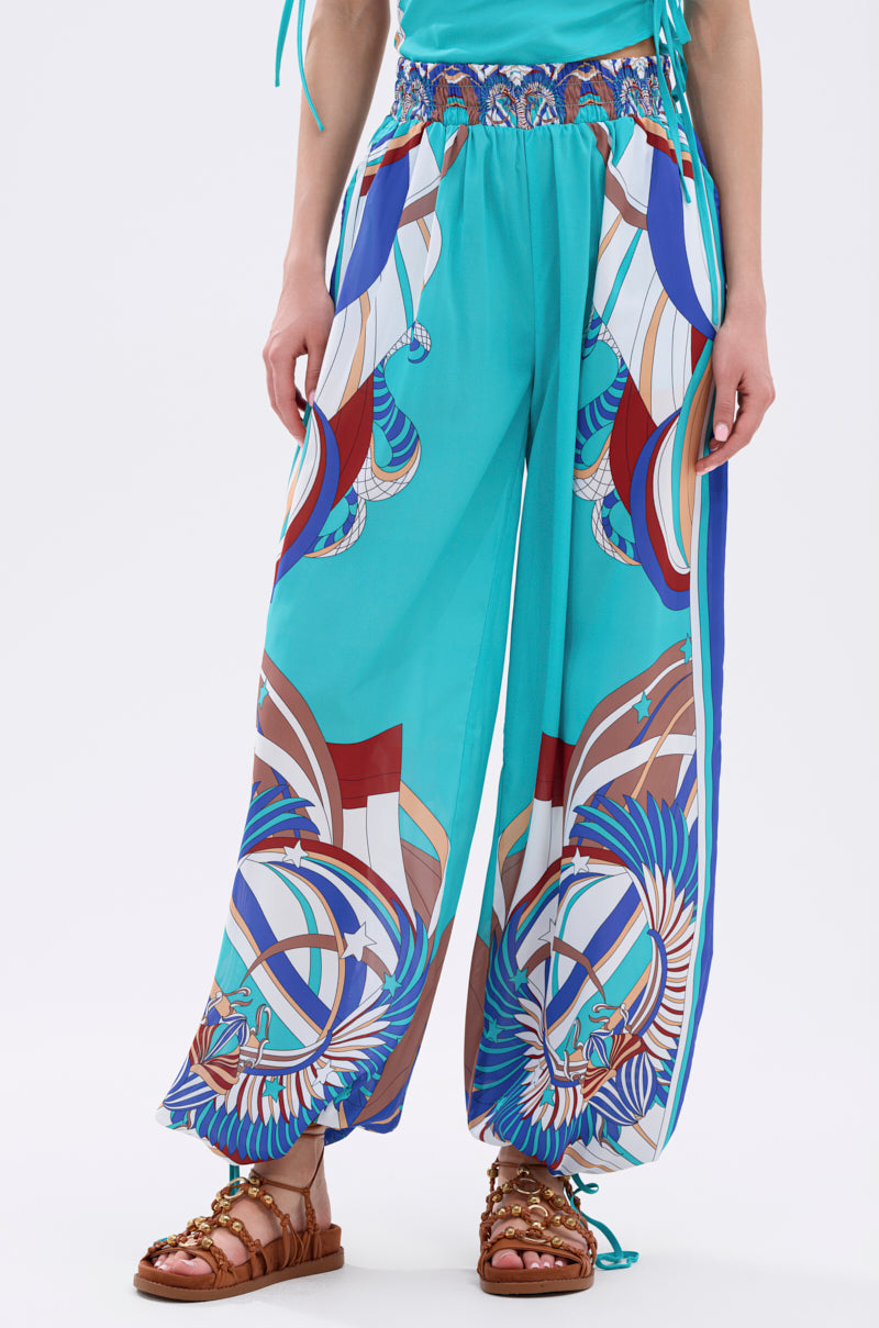 A person wears the ASHTIN PRINTED PALAZZO PANT with colorful geometric and abstract motifs, paired with brown strappy sandals. Only the lower half is shown against a plain background, capturing a chic resortwear look. 4