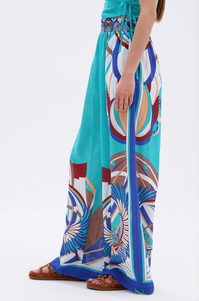 A woman stands sideways wearing the ASHTIN PRINTED PALAZZO PANT in bright turquoise with colorful geometric and abstract motifs, paired with brown woven sandals. Only her body from shoulder to foot is visible—a chic resortwear statement. 5