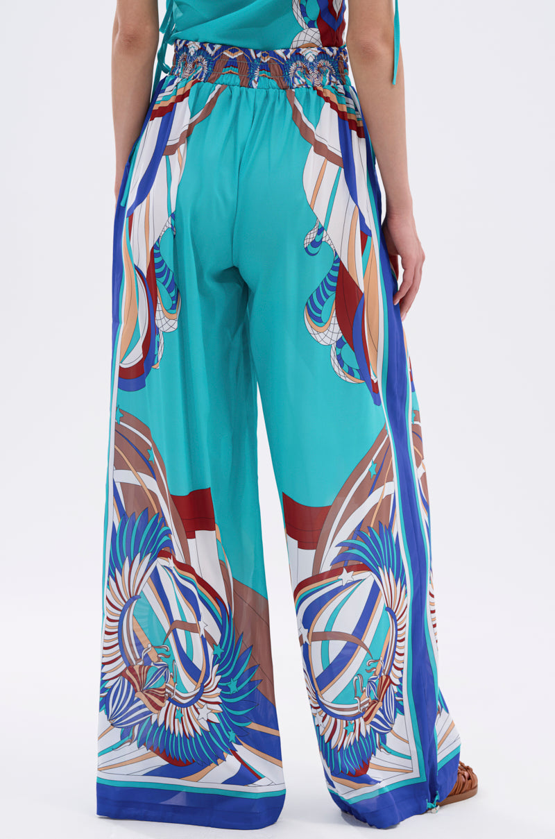A person models the ASHTIN PRINTED PALAZZO PANT, featuring loose teal fabric with a vibrant abstract bird-like print in blue, brown, white, and red. The back view highlights the flowy resortwear style and colorful design. 6