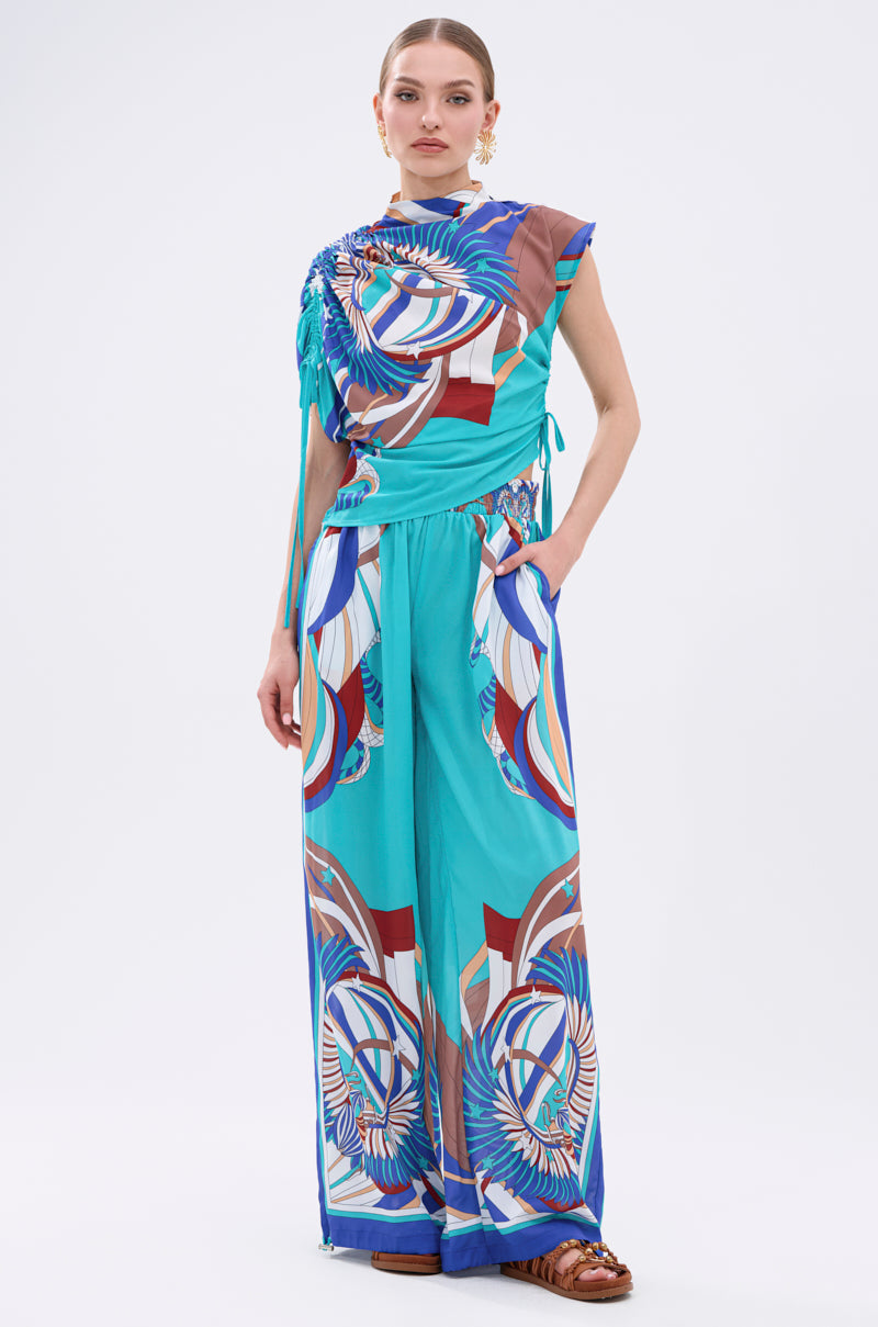 A woman stands facing forward, wearing the ASHTIN PRINTED PALAZZO PANT set with an abstract motif in turquoise, blue, red, and white. Her sleeveless top matches the palazzo pants. She has her hair pulled back and wears brown sandals. 7