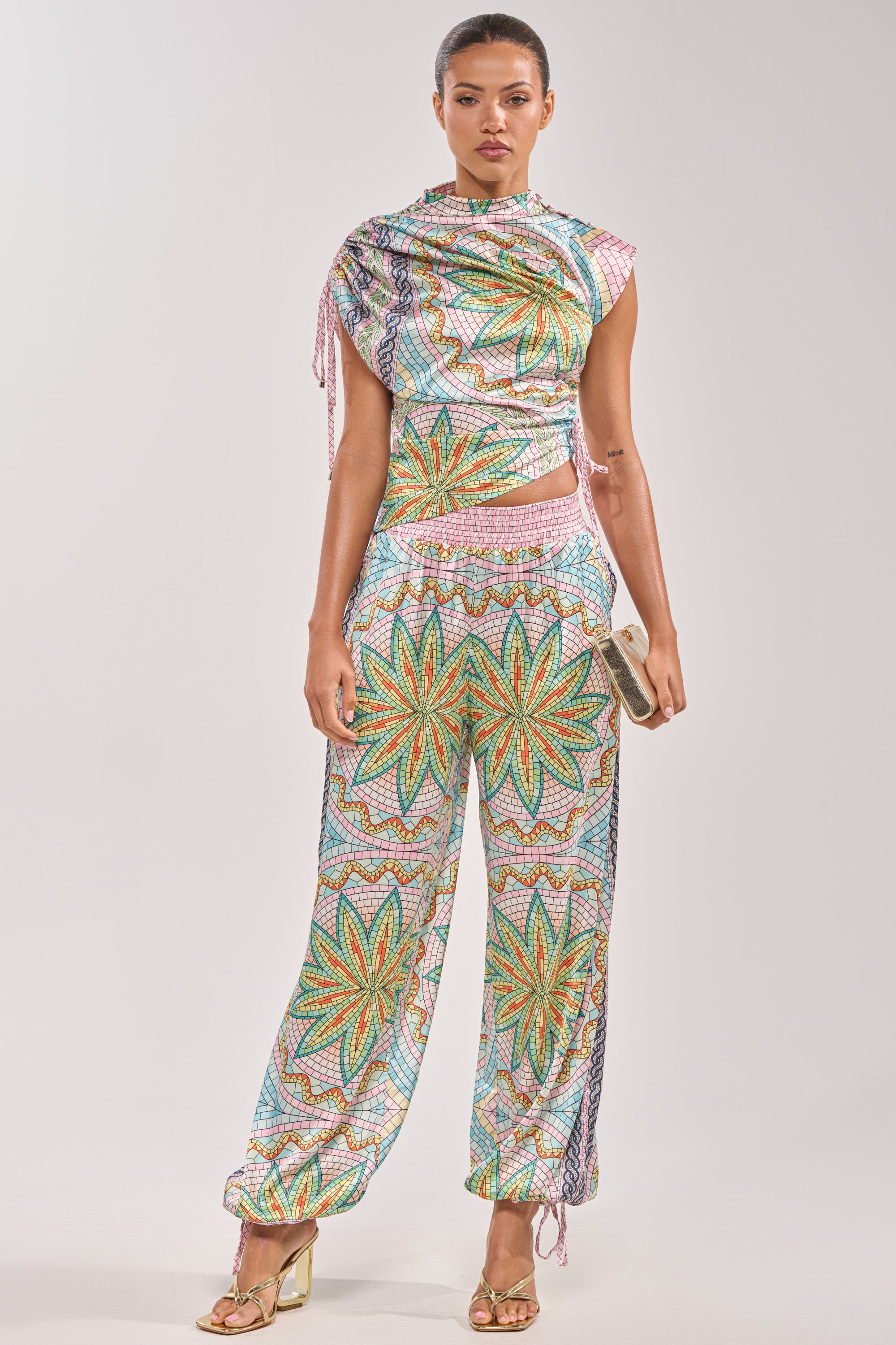 A woman wears the "ALL DIFFERENT WAYS SATIN PALAZZO PANT," a colorful, patterned wide-leg pant with green, orange, and yellow starburst designs—paired with a draped top for chic summer party style. She accessorizes with a clutch and gold heels. 0