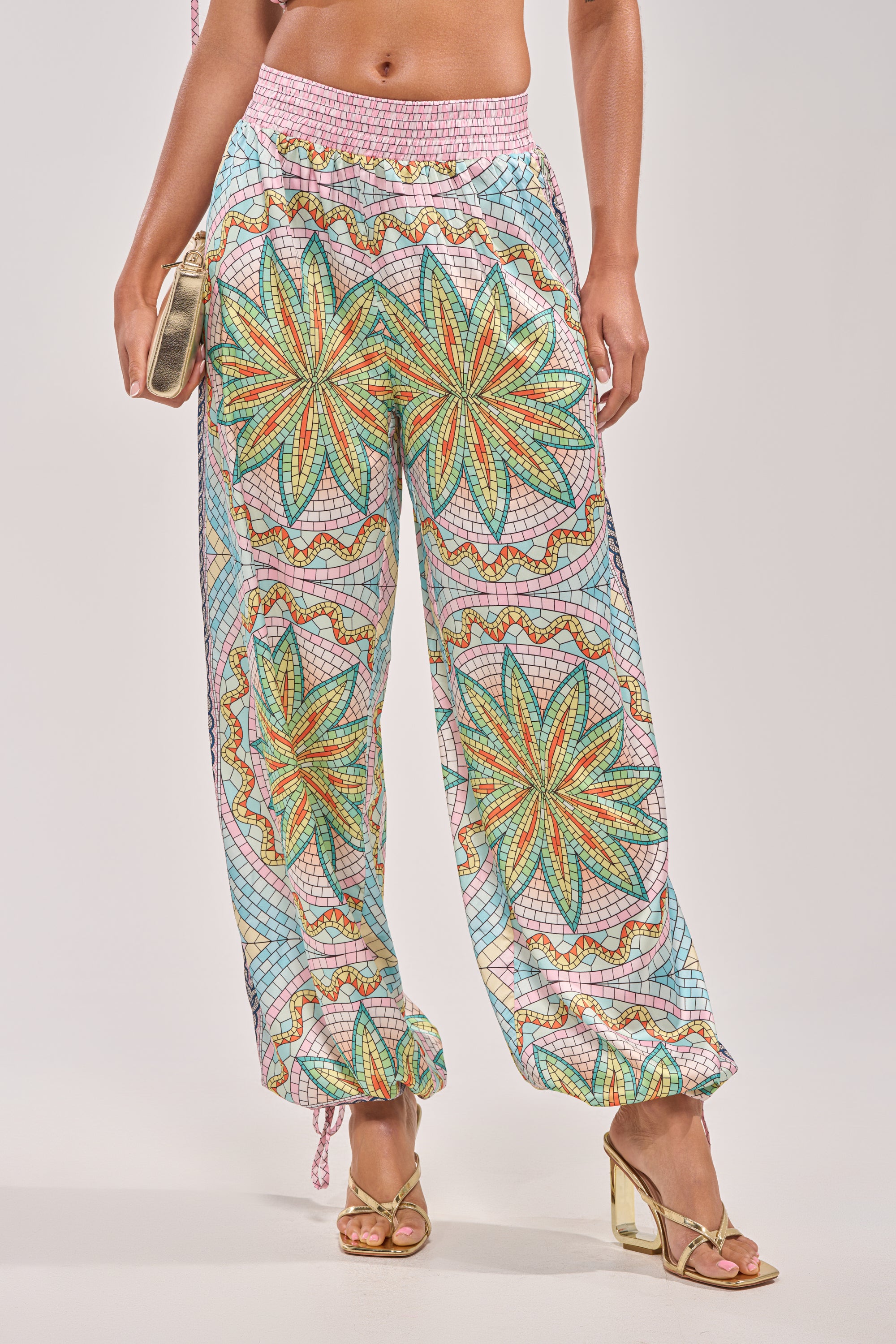 A wearer showcases the ALL DIFFERENT WAYS SATIN PALAZZO PANT—colorful wide-leg pants with starburst designs—paired with gold heels and a beige clutch, shown from shoulders down against a plain background; ideal for summer parties. 1