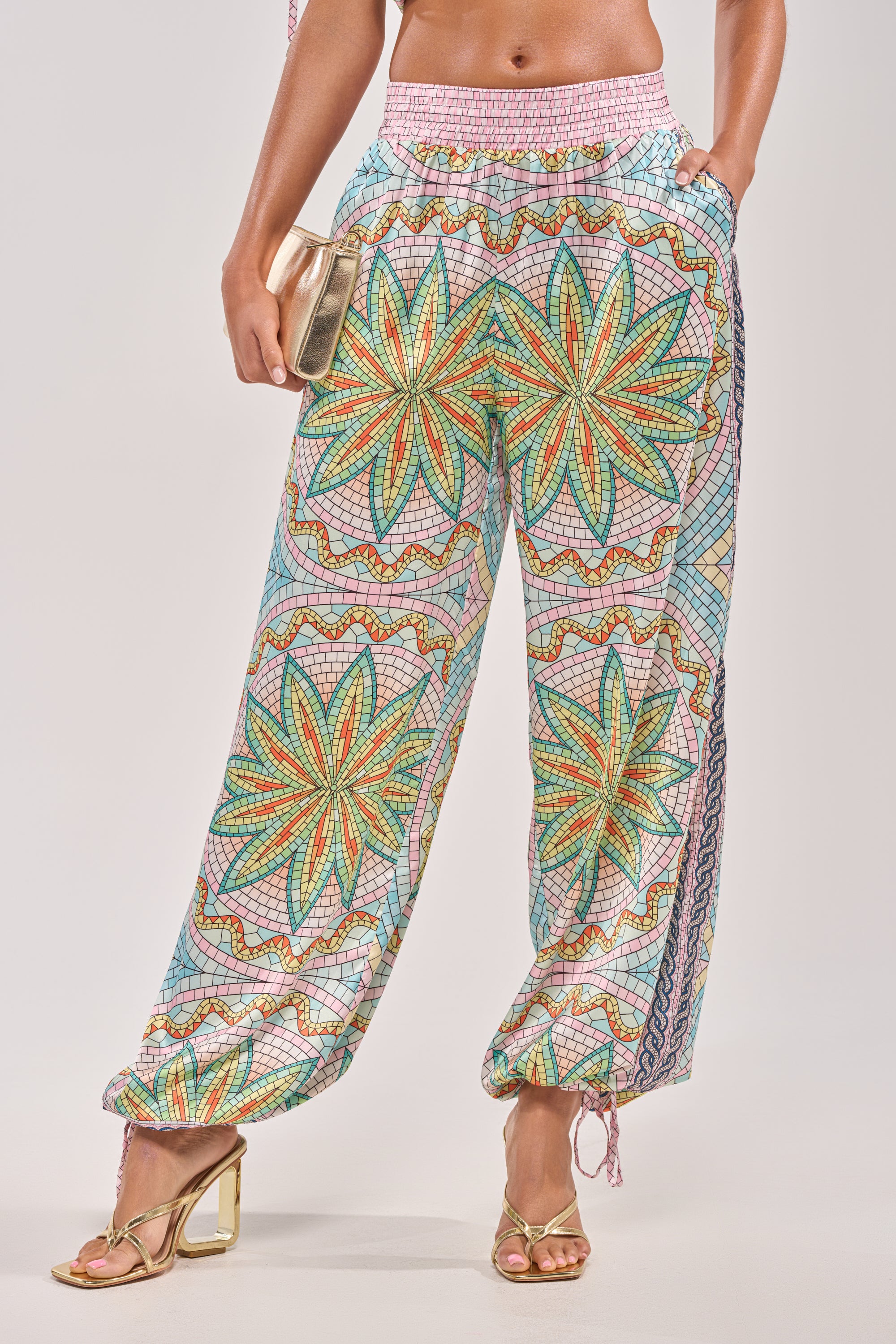 A person wearing the ALL DIFFERENT WAYS SATIN PALAZZO PANT with a vibrant floral design, paired with gold heeled sandals, and holding a small gold clutch bag. The background is plain and light-colored. 2