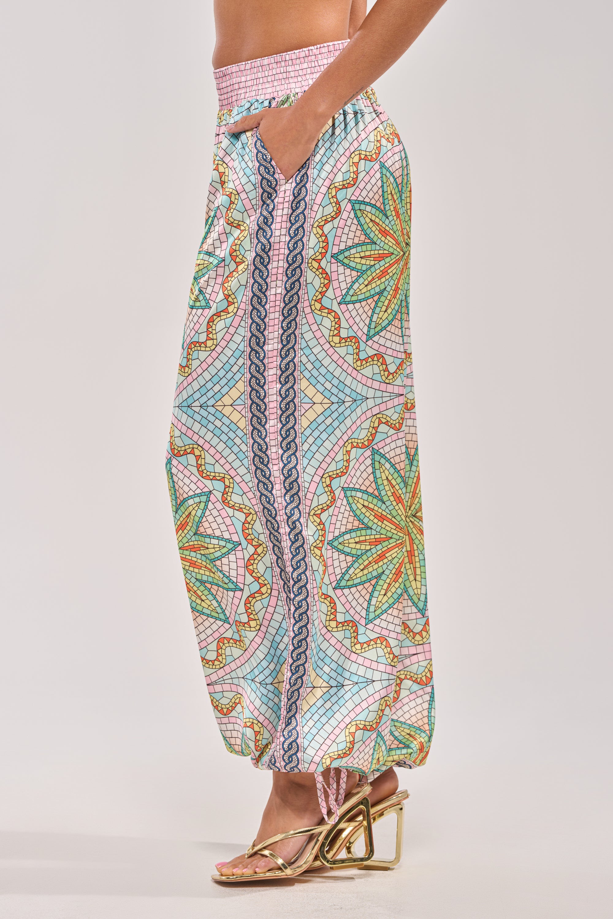 A person wears the ALL DIFFERENT WAYS SATIN PALAZZO PANT, showcasing wide legs, a high waist, elastic cuffs, and geometric and floral prints in green, yellow, red, and blue. They pair it with gold strappy sandals featuring a unique heel design. 3