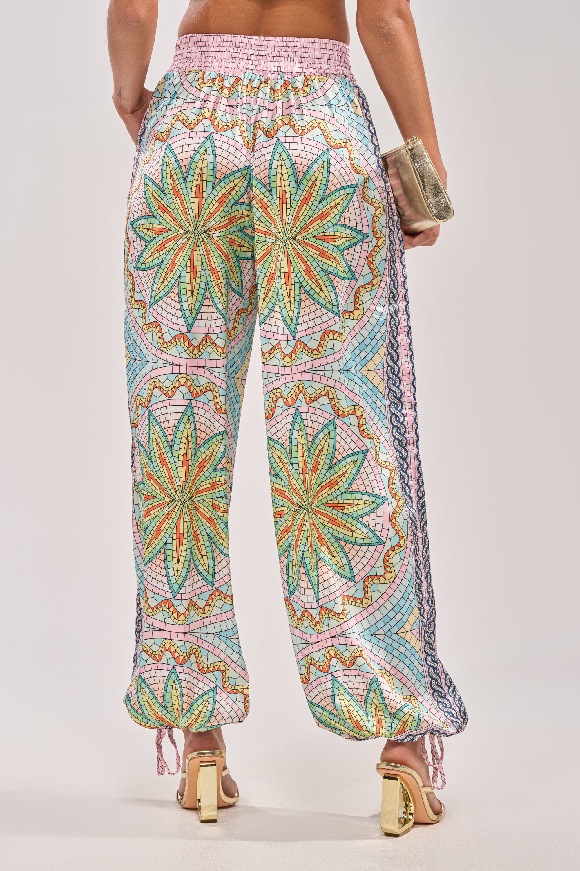 A person wearing the ALL DIFFERENT WAYS SATIN PALAZZO PANT, featuring a bold geometric floral print in green, orange, and blue, paired with gold heeled sandals and holding a small metallic clutch. 4