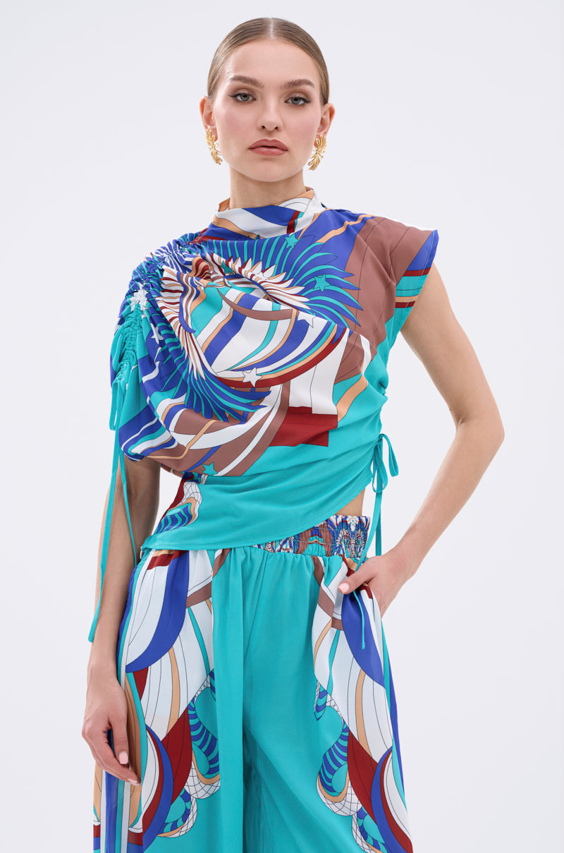 A woman stands confidently in the ASHTIN PRINTED BLOUSE, a vibrant multicolor, asymmetrical top paired with matching turquoise geometric pants. She poses with one hand in her pocket against a plain white background. 0