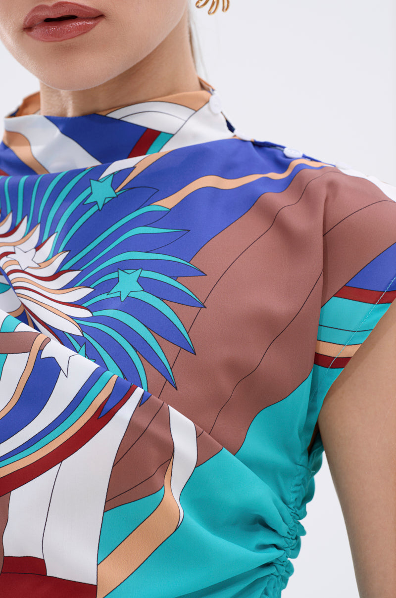 A close-up shows a woman wearing the ASHTIN PRINTED BLOUSE with a bold multicolor abstract print in blue, brown, turquoise, and white. Only her lips, chin, and shoulder are visible; she wears gold earrings for effortless resortwear chic. 10