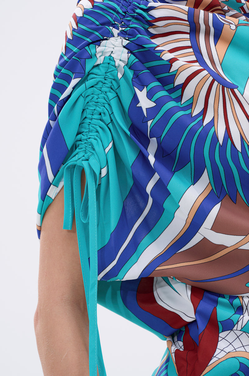 Close-up of the ASHTIN PRINTED BLOUSE, an asymmetrical multicolor top featuring a bold abstract pattern in blue, teal, brown, white, and red. Ruched resortwear fabric and drawstring sleeve ties create a stylish gathered look. 11