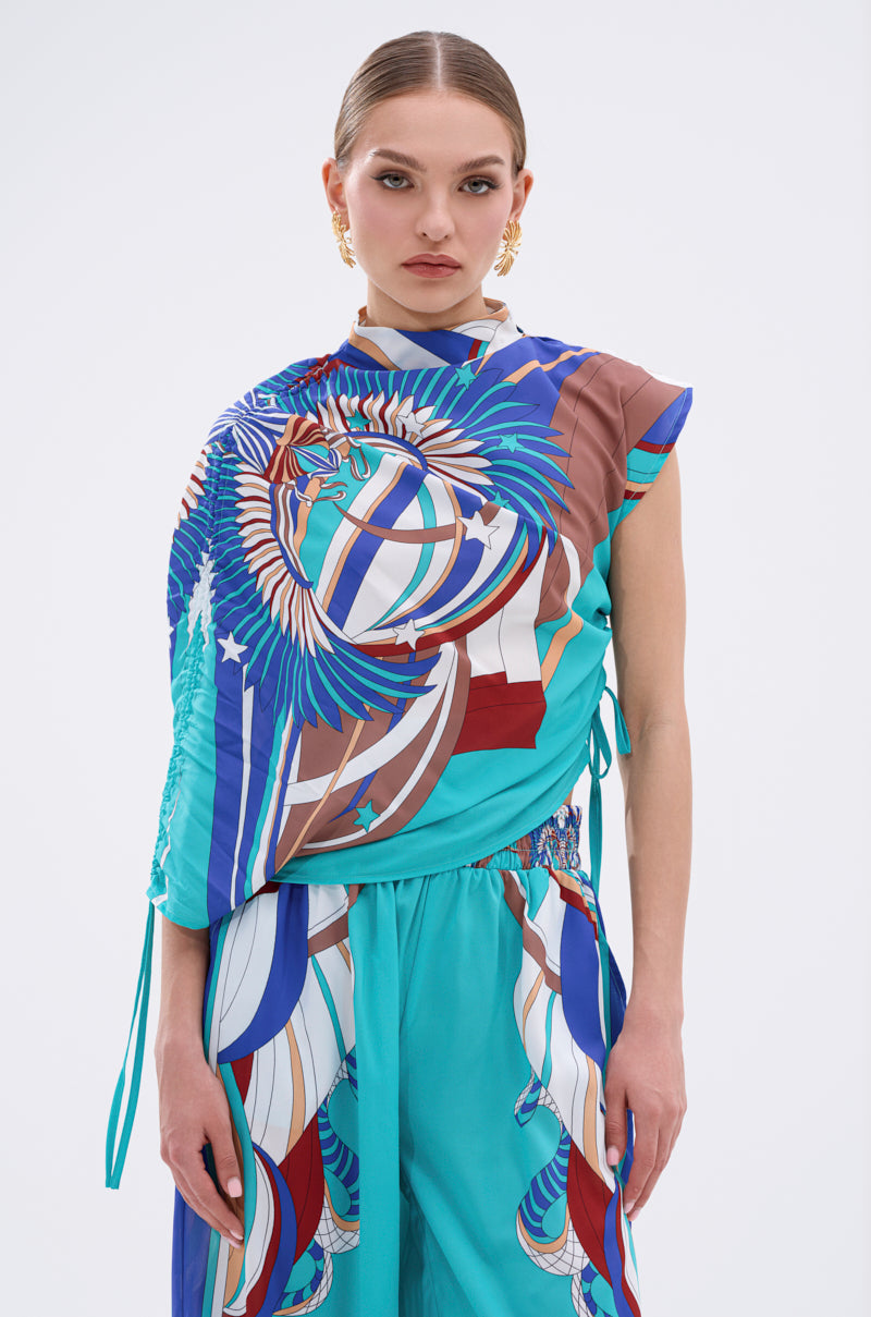 A woman with slicked-back hair wears the ASHTIN PRINTED BLOUSE, a vibrant, multicolor asymmetrical top with an abstract eagle and flag print and gold earrings. She stands against a plain white background, looking directly at the camera. 1
