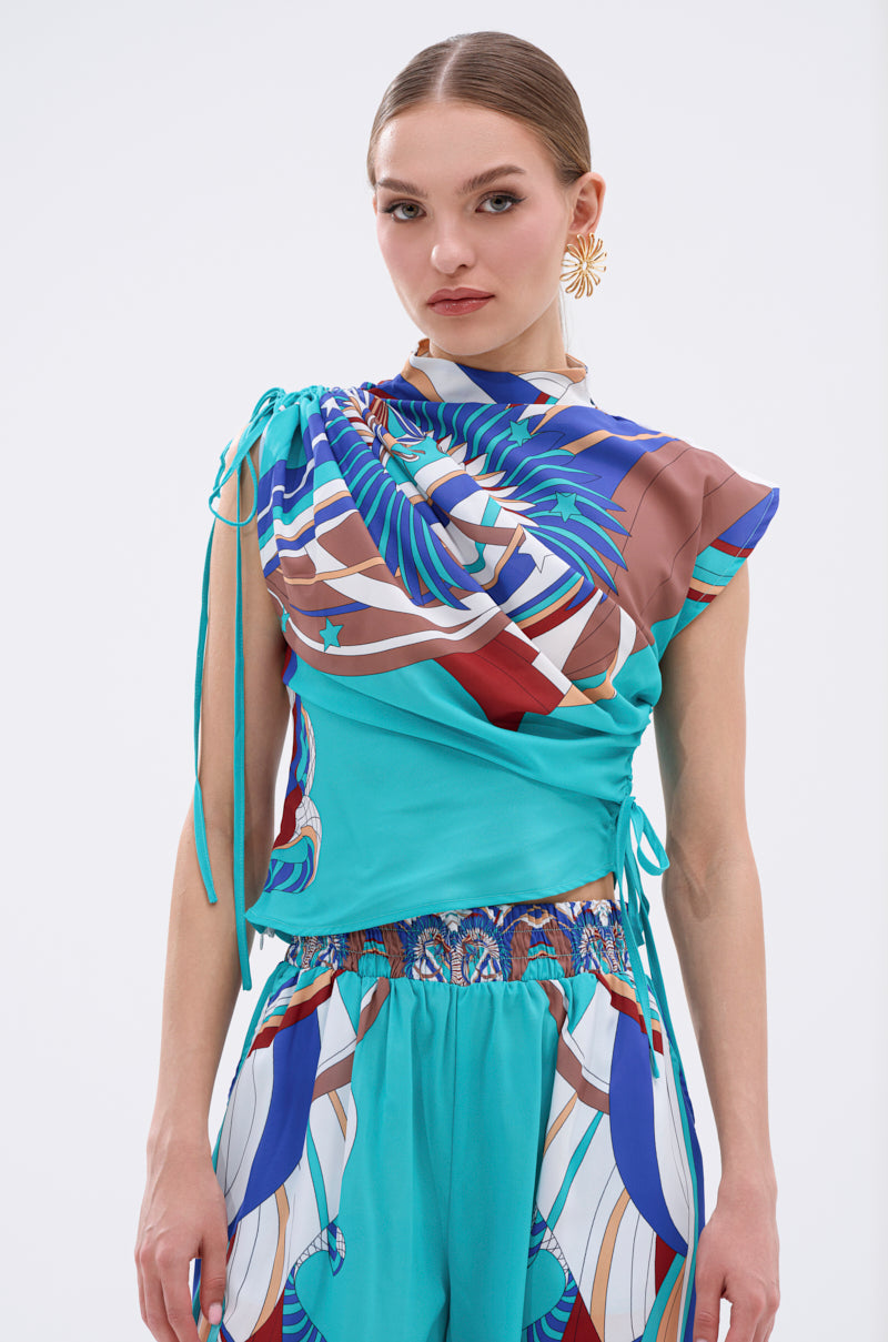 A woman stands against a plain light background wearing the ASHTIN PRINTED BLOUSE in a vibrant multicolor print with drawstring details, paired with matching high-waisted pants in teal, brown, blue, and white. 2