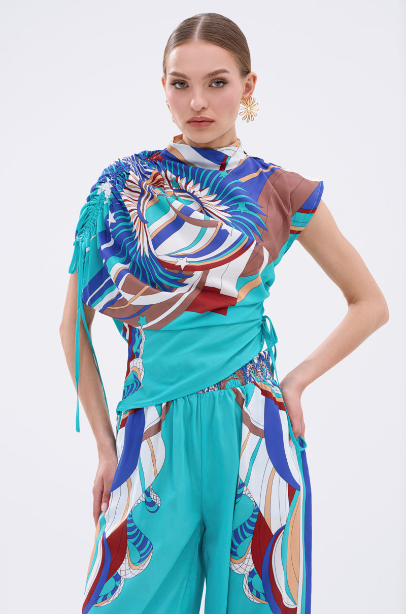 A woman poses confidently in the ASHTIN PRINTED BLOUSE, a vibrant multicolor resortwear top with abstract blue, white, and brown designs. The asymmetrical blouse features ruched and draped details on one shoulder. She wears gold earrings and has slicked-back hair. 3