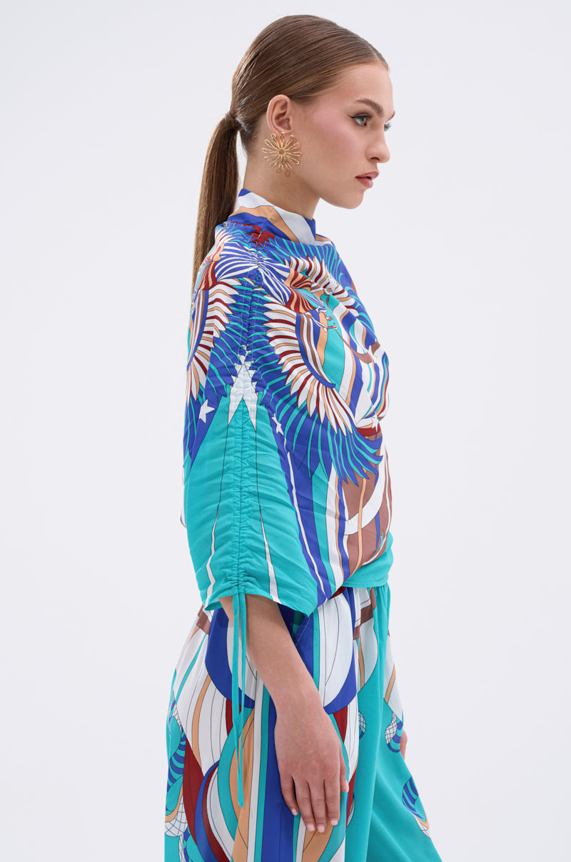 A woman with straight brown hair in a low ponytail wears the ASHTIN PRINTED BLOUSE with a vibrant multicolor print and large floral earrings, showcasing effortless resortwear style against a plain white background. 5