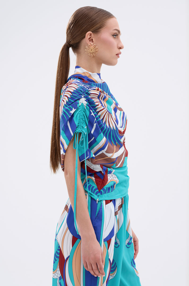 A woman with long straight brown hair in a low ponytail models the ASHTIN PRINTED BLOUSE featuring a multicolor graphic print with blue, turquoise, and brown swirls. She wears gold flower-shaped earrings and stands in profile against a white background. 6
