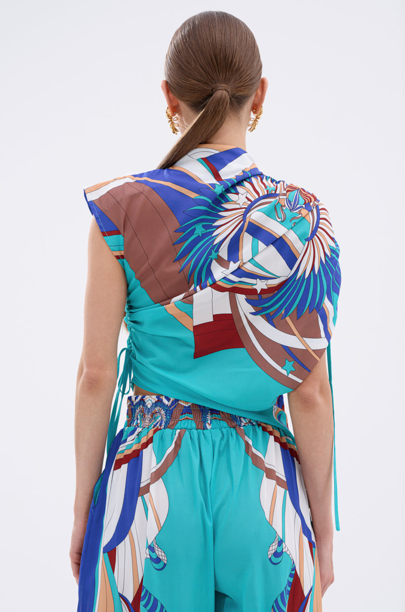 A woman with straight brown hair in a low ponytail is seen from behind, wearing the ASHTIN PRINTED BLOUSE with multicolor graphics, coordinated turquoise abstract-print pants, and gold earrings. 7