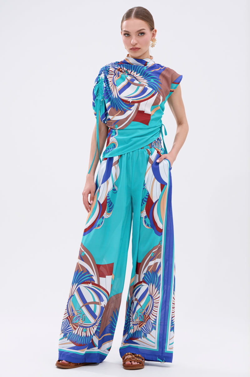 A woman stands against a white background in the ASHTIN PRINTED BLOUSE with matching multicolor graphic wide-leg pants—an ideal resortwear look in blue, teal, brown, and white tones, styled with chic brown sandals. 8