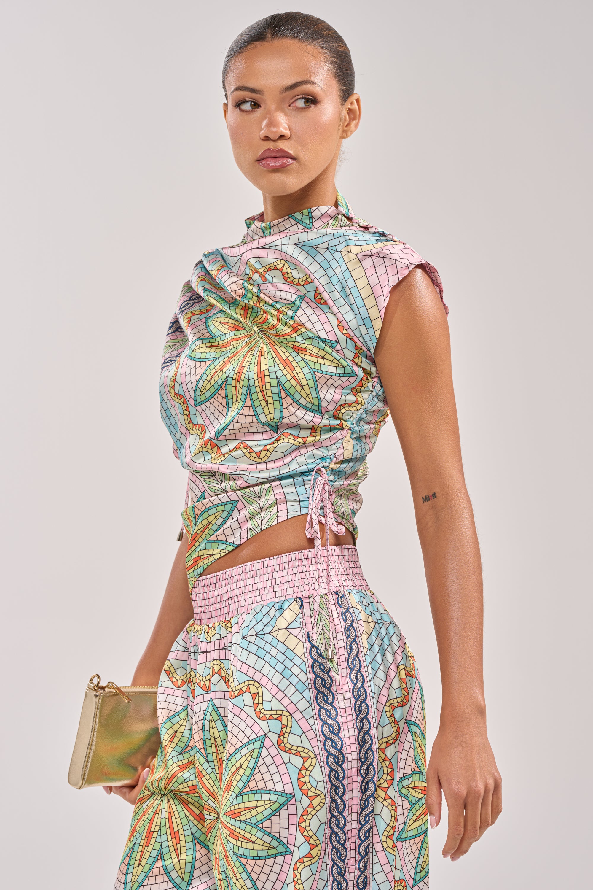 A woman with pulled-back hair and a neutral look wears the ALL DIFFERENT WAYS SATIN BLOUSE in a colorful geometric pattern, styled as a two-piece outfit. She looks to the side, holding a small gold clutch. 2