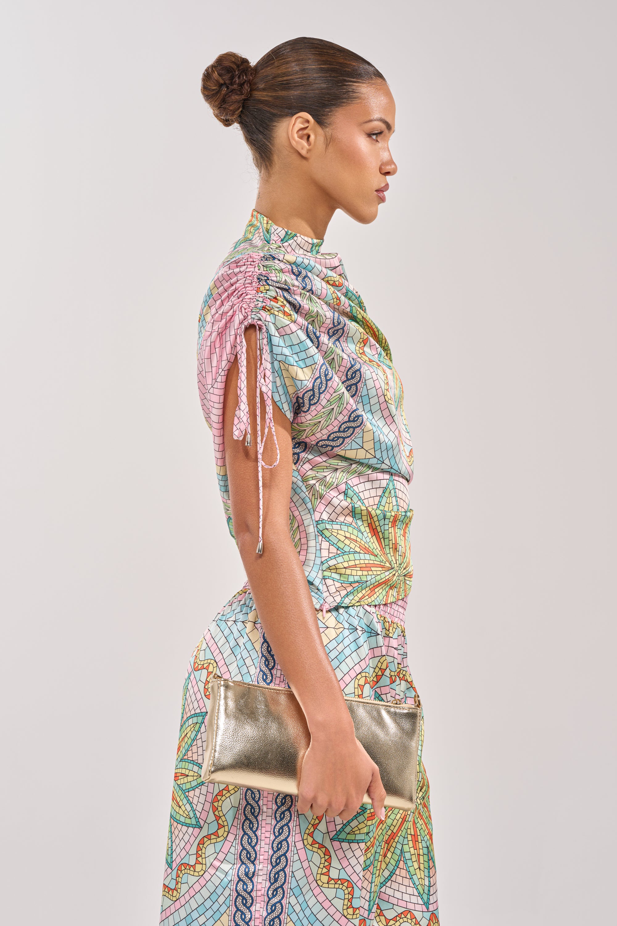 A woman with a neat bun stands in profile against a plain background, wearing the ALL DIFFERENT WAYS SATIN BLOUSE—a pink mosaic top with ruched sleeves. She holds a shiny gold clutch, creating an elegant summer party look. 3